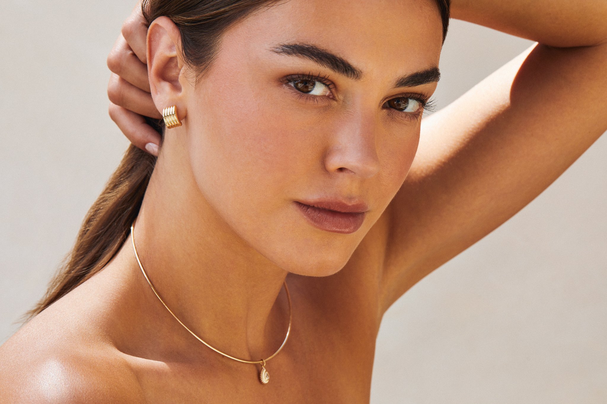A woman with brown hair and brown eyes wearing gold jewelry, including a necklace, earrings, and a ring. She has one arm raised and her hand near her ear, with a neutral background.