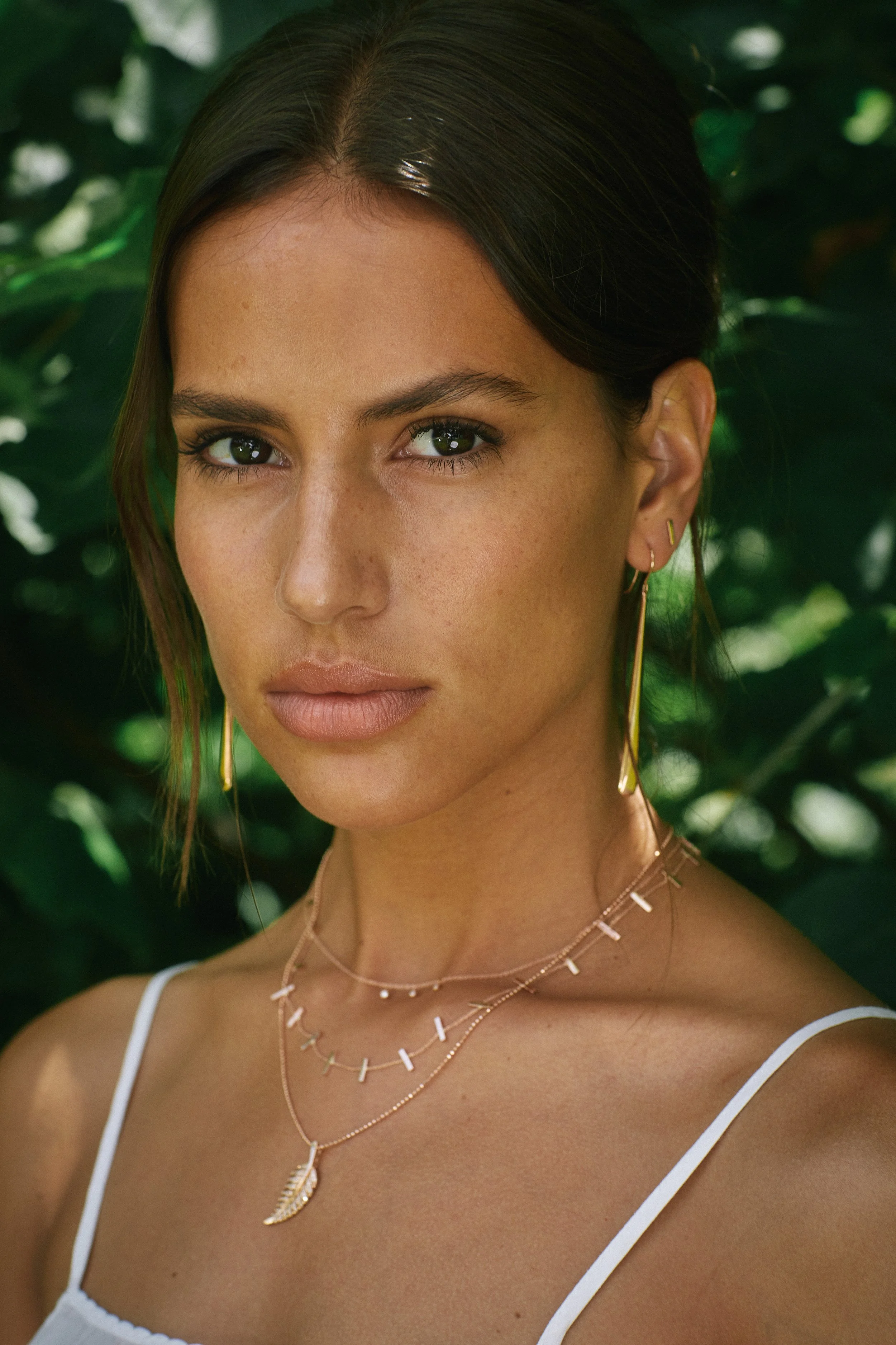 Close-up of a woman with dark hair, wearing layered necklaces and earrings, standing outdoors with lush green foliage in the background.