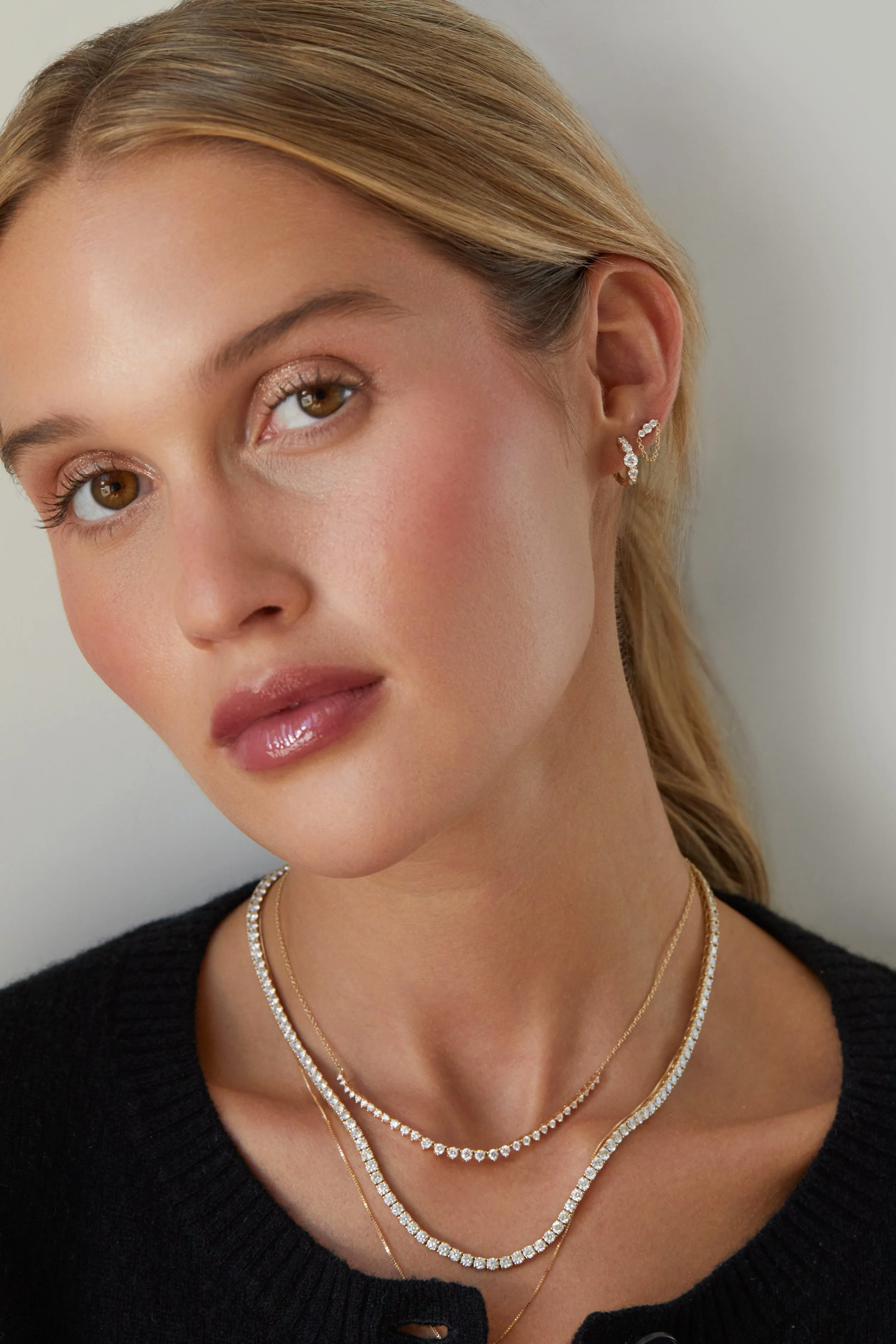 Close-up of a woman with blonde hair wearing multiple layered diamond necklaces, diamond earrings, and a black top against a neutral background.