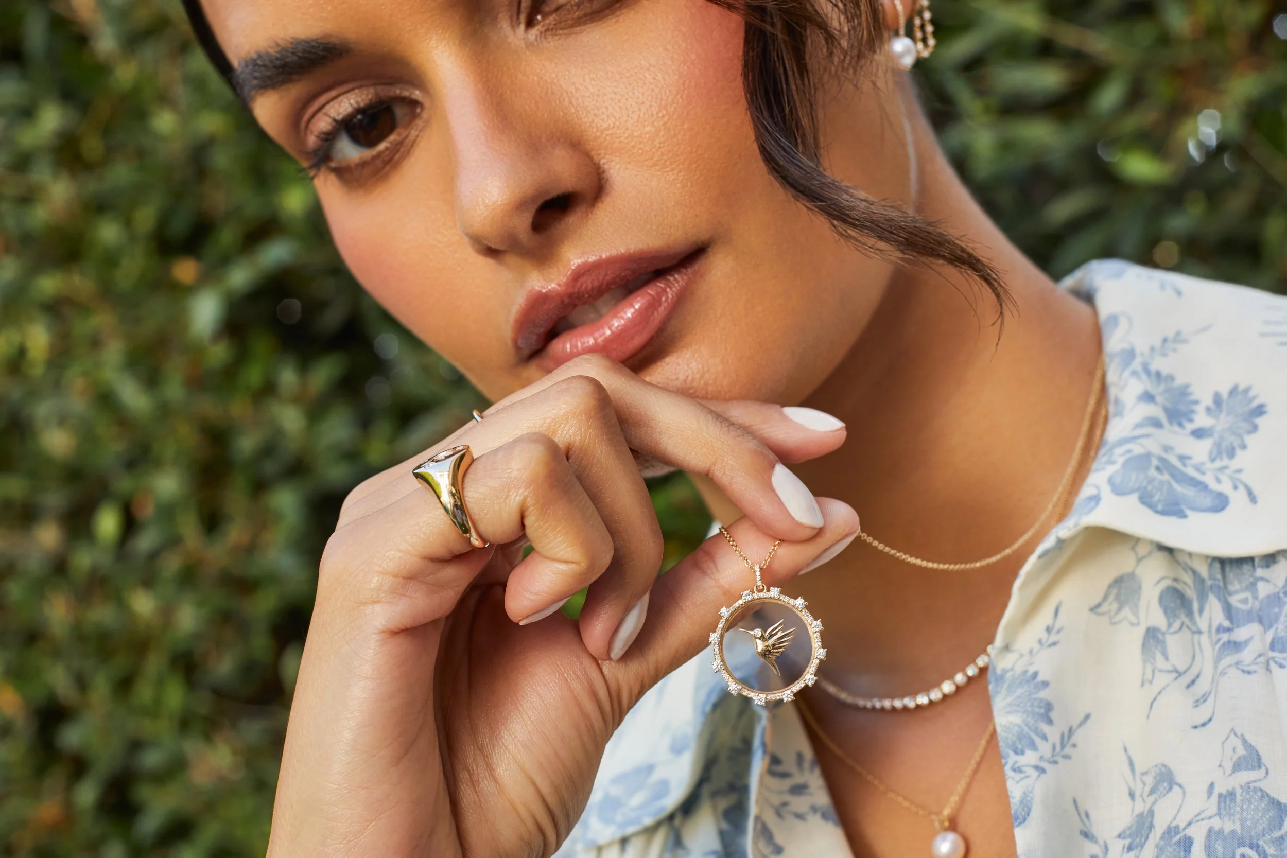 Close-up of a woman holding a round pendant necklace adorned with a dove silhouette, wearing layered pearl and gold necklaces, with a gold ring on her finger, against a blurred green outdoor background.