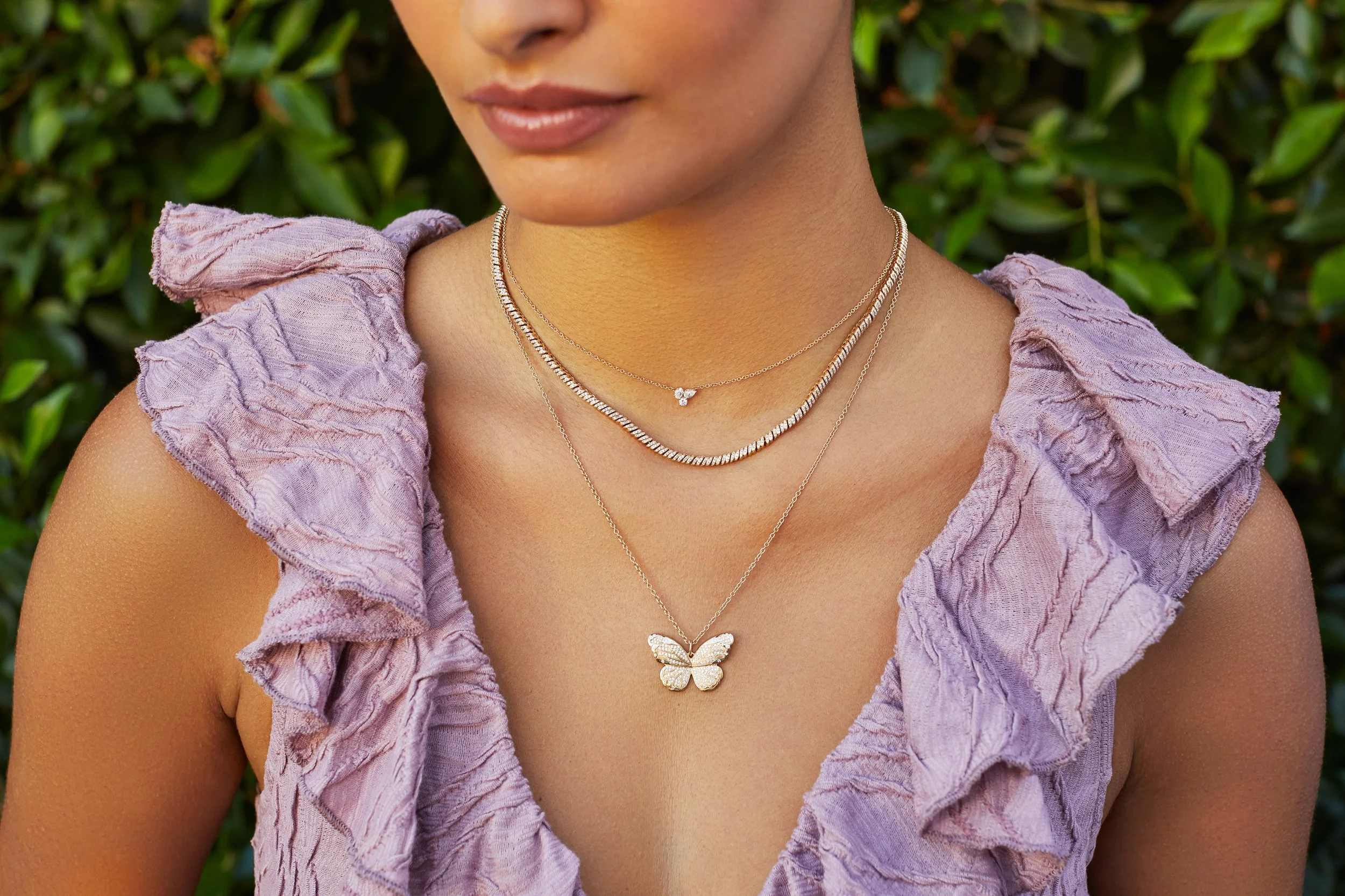 Close-up of a woman wearing layered necklaces, including one with a butterfly pendant, with a purple ruffled top and green foliage in the background.