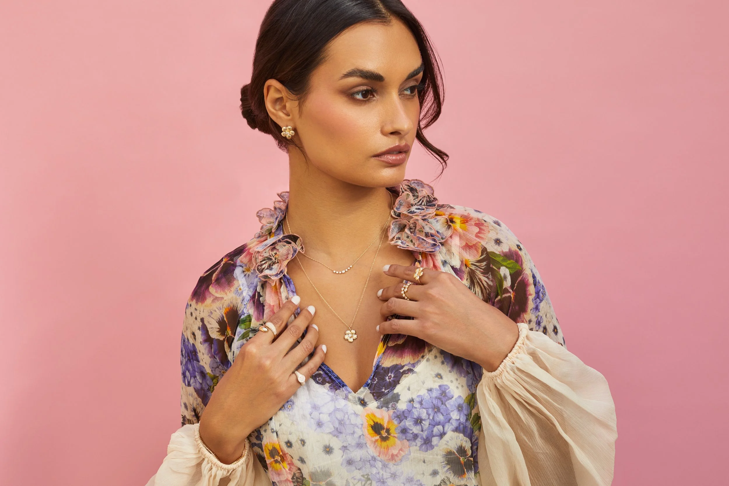 A woman wearing a floral blouse and jewelry against a pink background.
