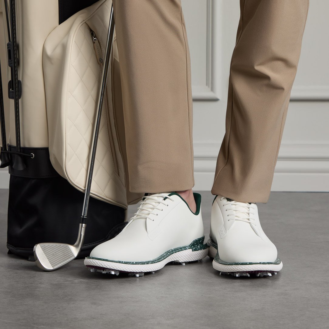 Close-up of a person wearing beige golf pants and white golf shoes, standing next to a golf bag with clubs.