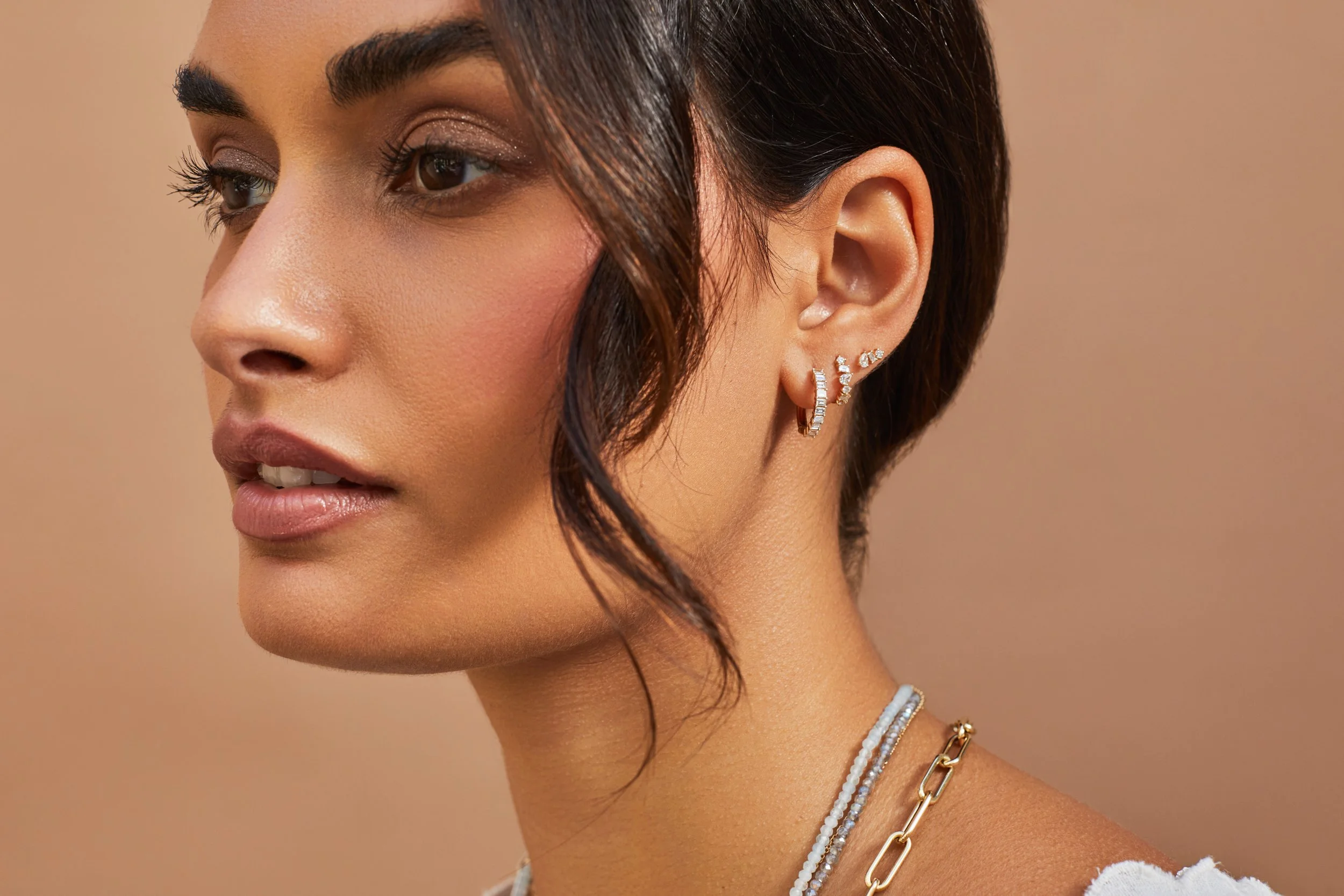 A woman with short dark hair wearing multiple earrings, including a diamond hoop and smaller diamond studs, and layered necklaces against a beige background.