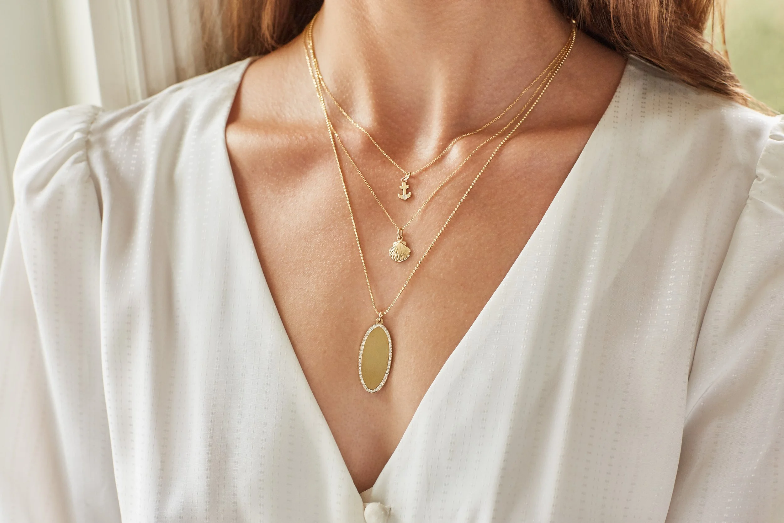 Close-up of a woman's neck and chest wearing layered gold necklaces with sea shell, anchor, and oval pendant, paired with a white blouse with puffed sleeves.