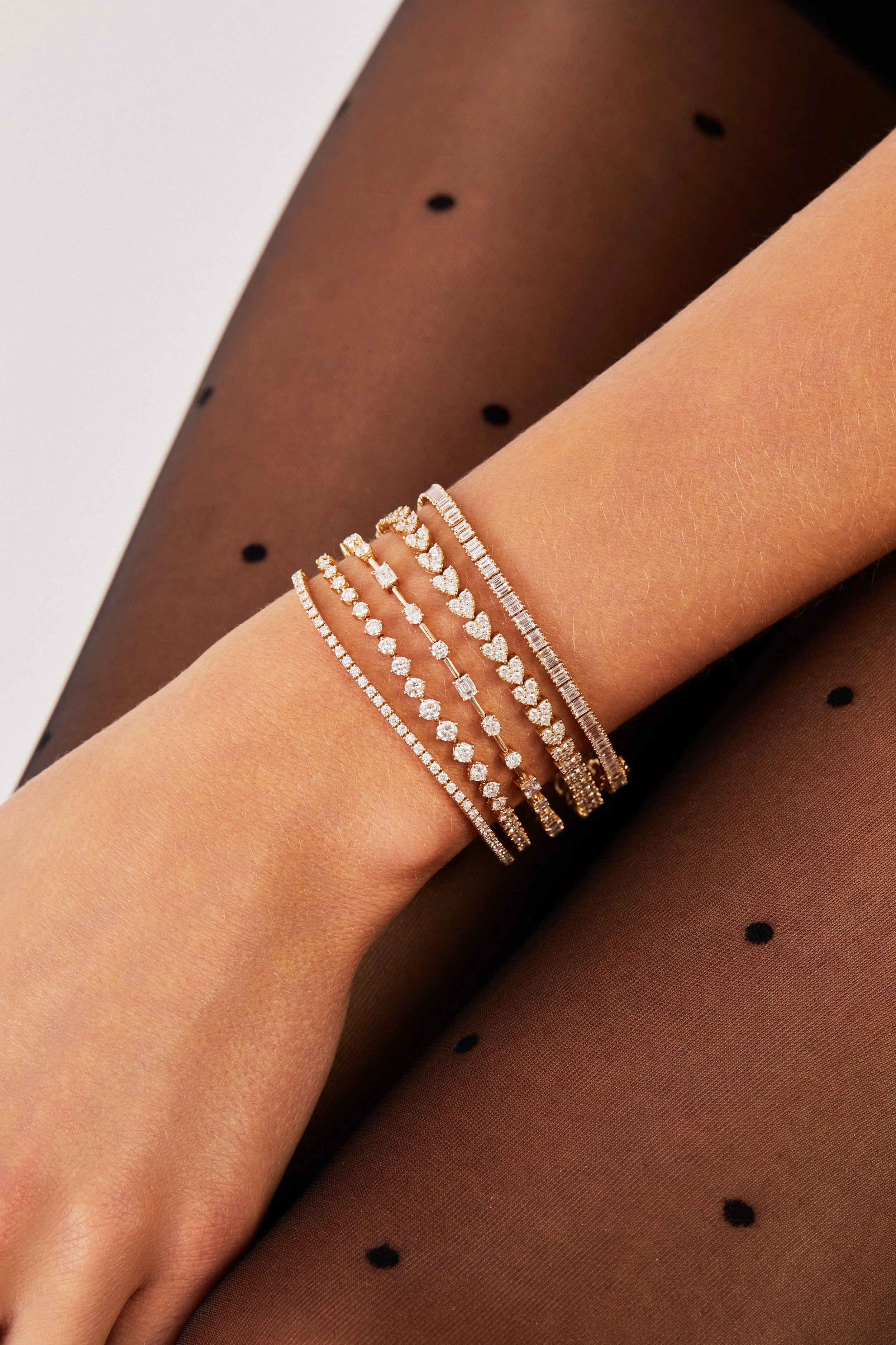 A person's arm wearing five different diamond bracelets, with sheer black polka dot tights visible on the leg.
