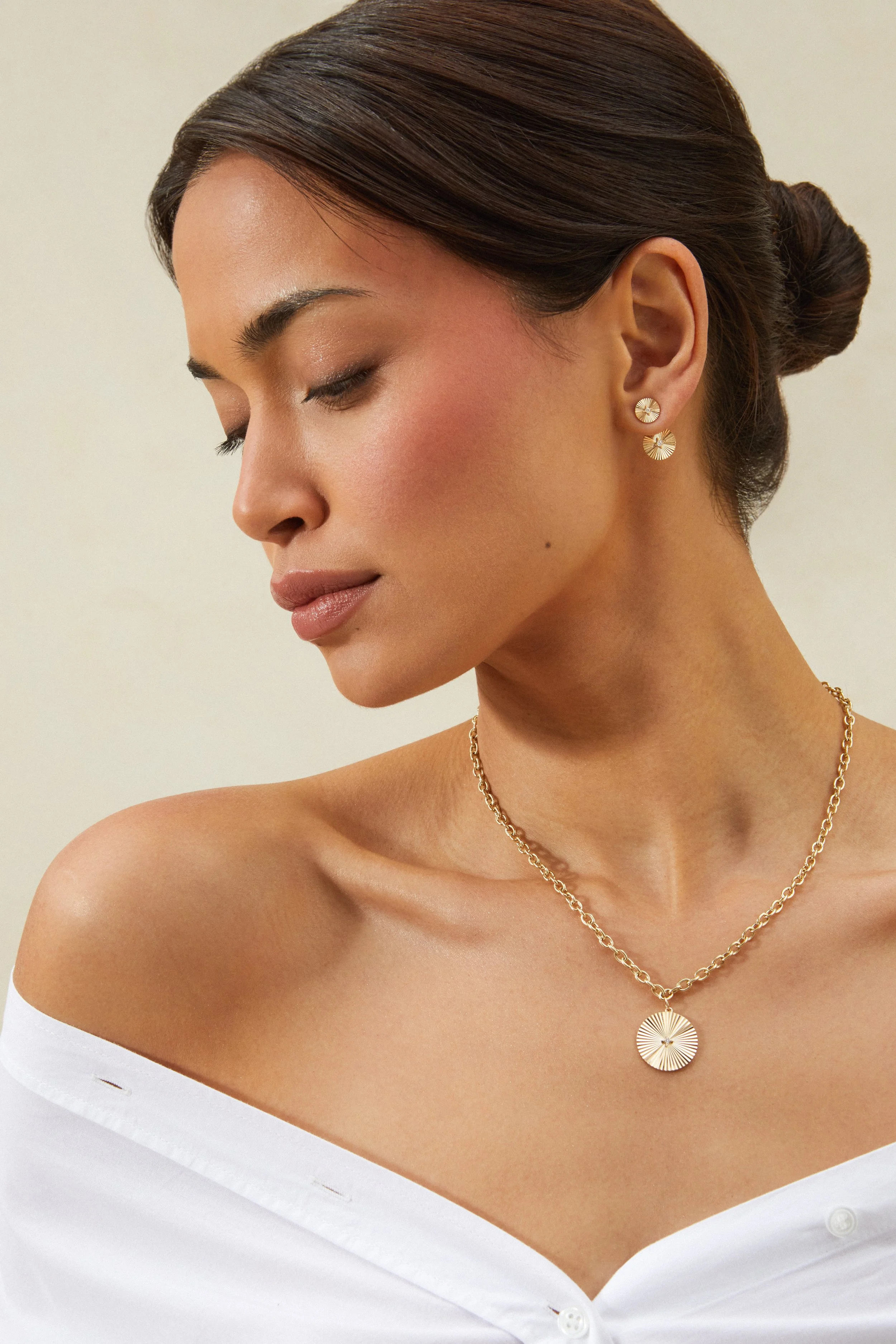 A woman with dark hair styled in a bun, wearing gold jewelry including earrings and a necklace, with her eyes closed and her head slightly tilted downward.