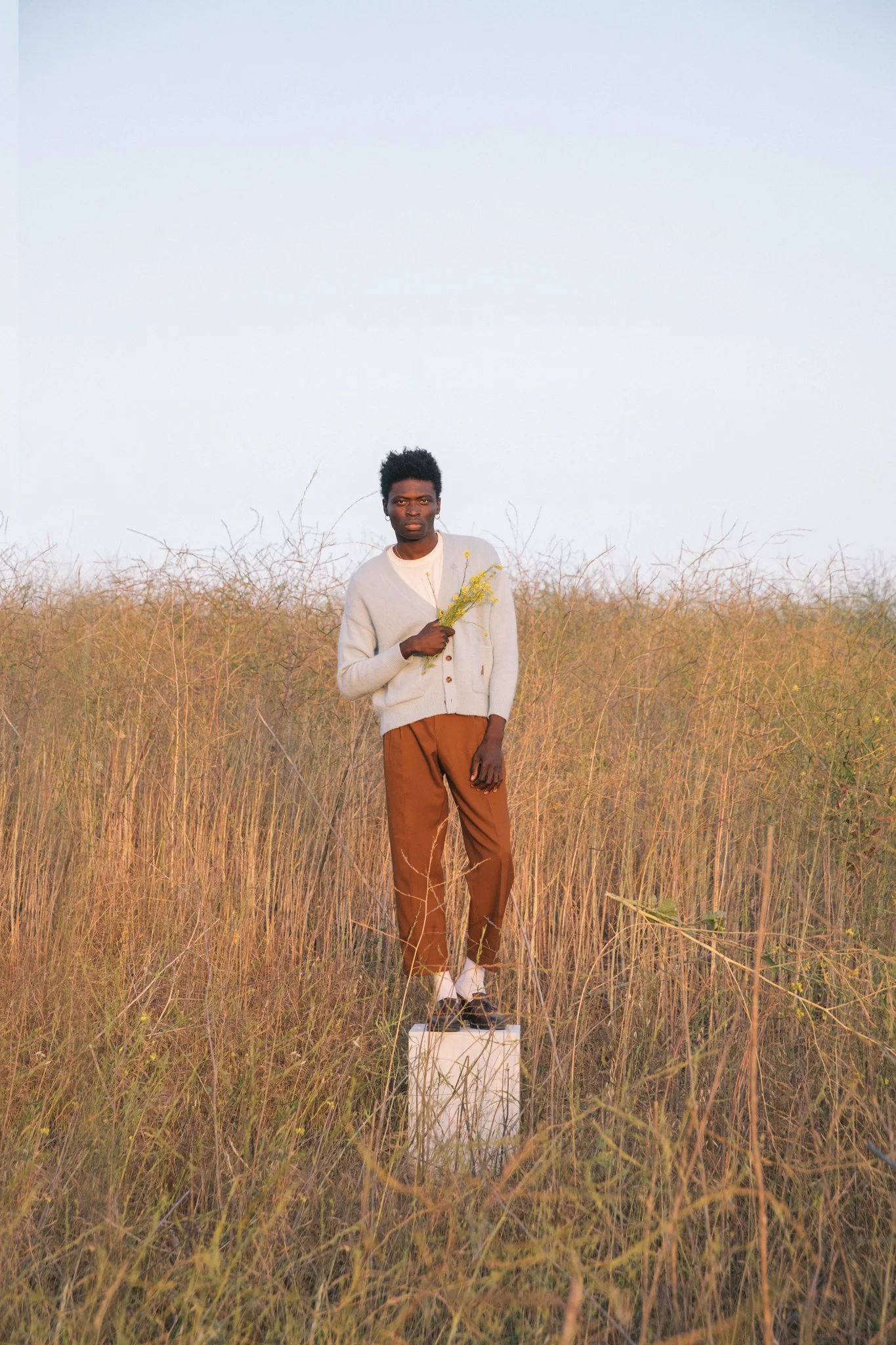 A young Black man standing on a white box in a field of tall dry grass, holding a small bouquet of yellow flowers, with a clear sky in the background.