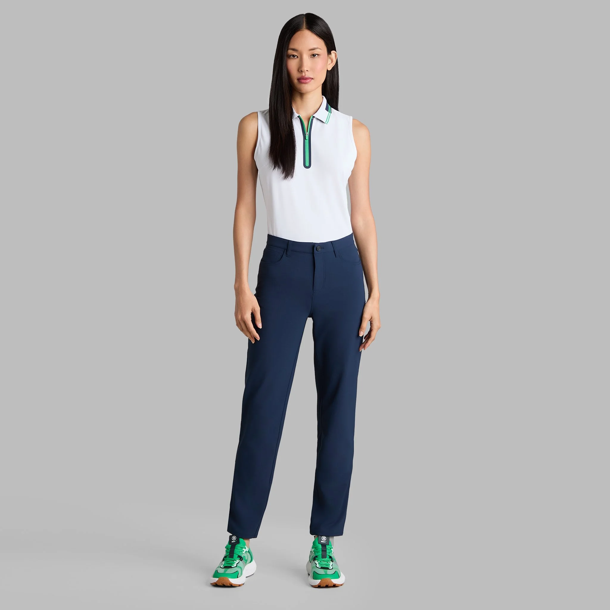 A woman wearing a sleeveless white sports top, navy blue pants, and green sneakers standing against a gray background.