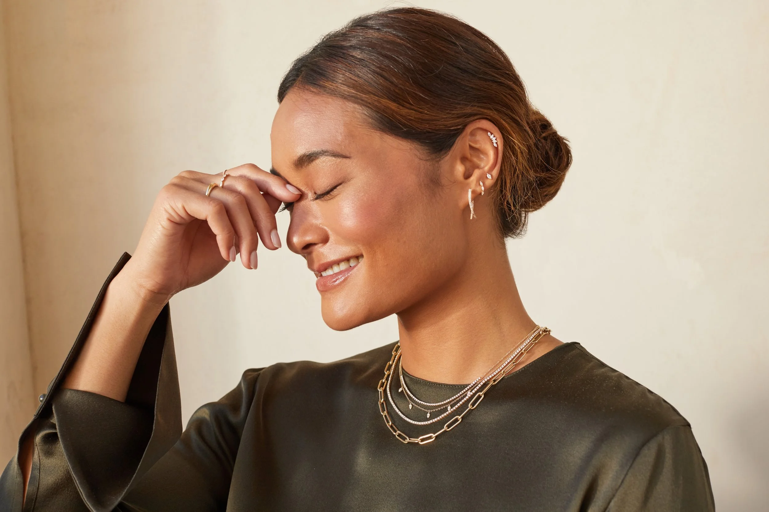 A woman with short brown hair in a bun smiling with her eyes closed, wearing multiple layered necklaces and earrings, dressed in a dark shiny top, standing against a plain beige background.