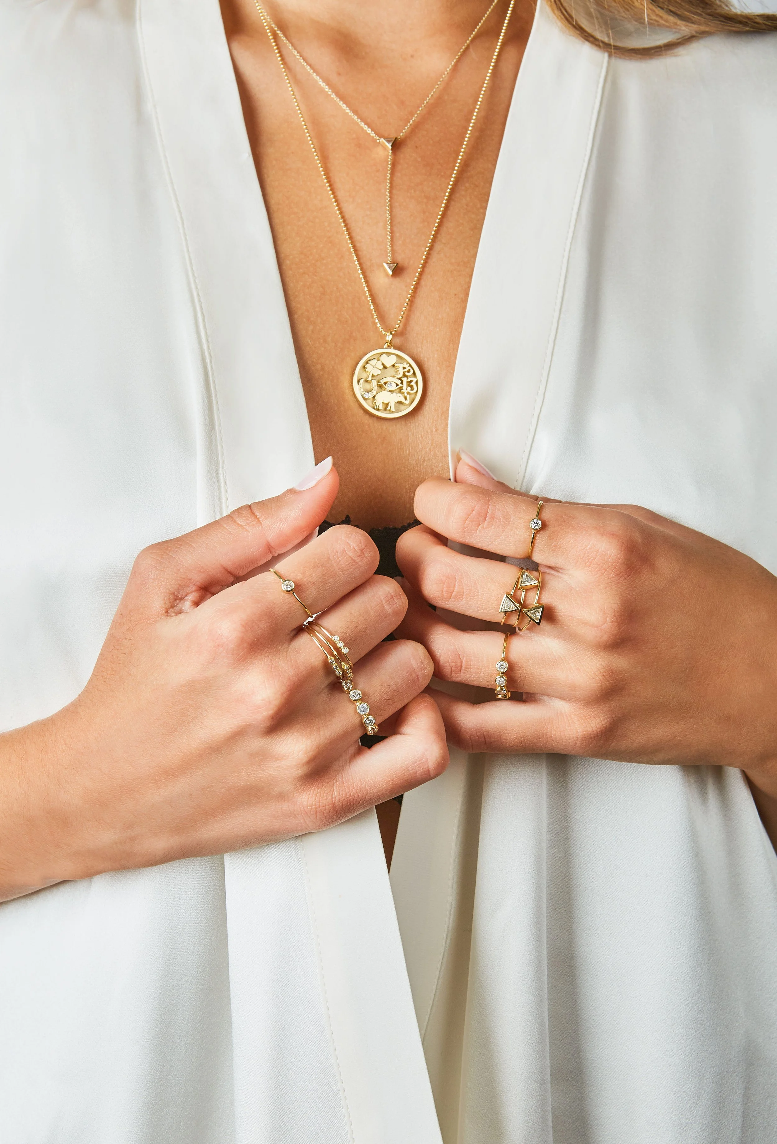 Close-up of a woman wearing layered gold necklaces, rings, and holding her hands together, showcasing jewelry.