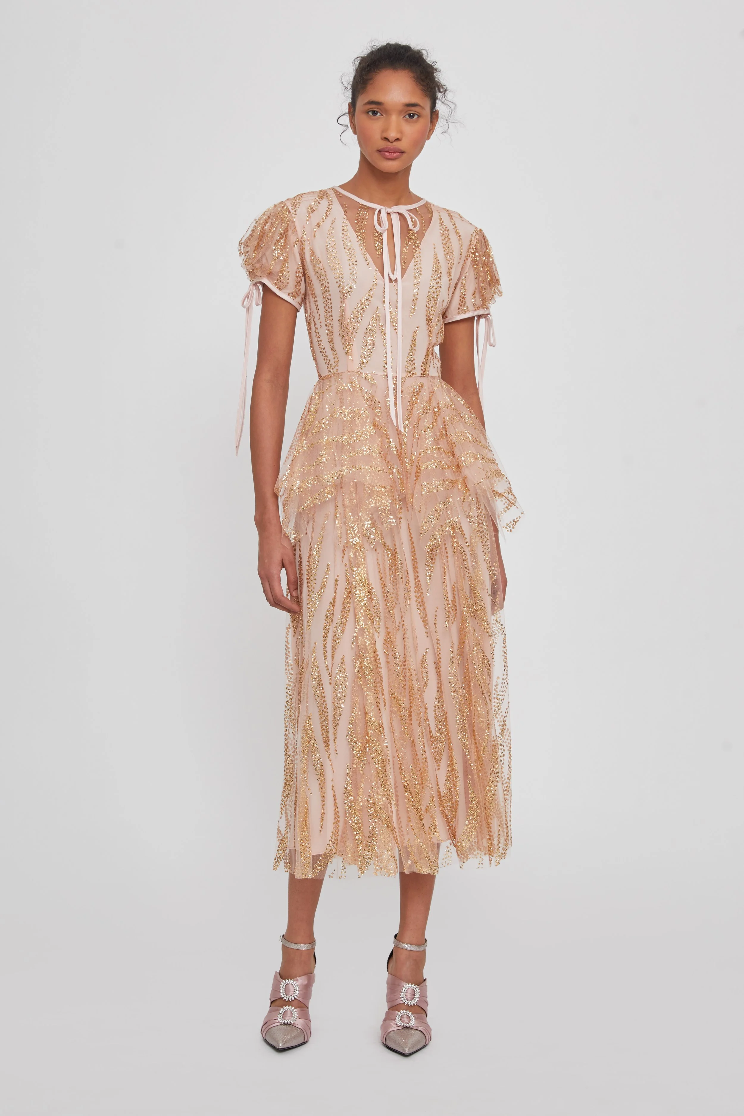 Model wearing a pink and gold embellished dress with layered ruffles and sheer fabric, paired with pink heels with decorative bows.