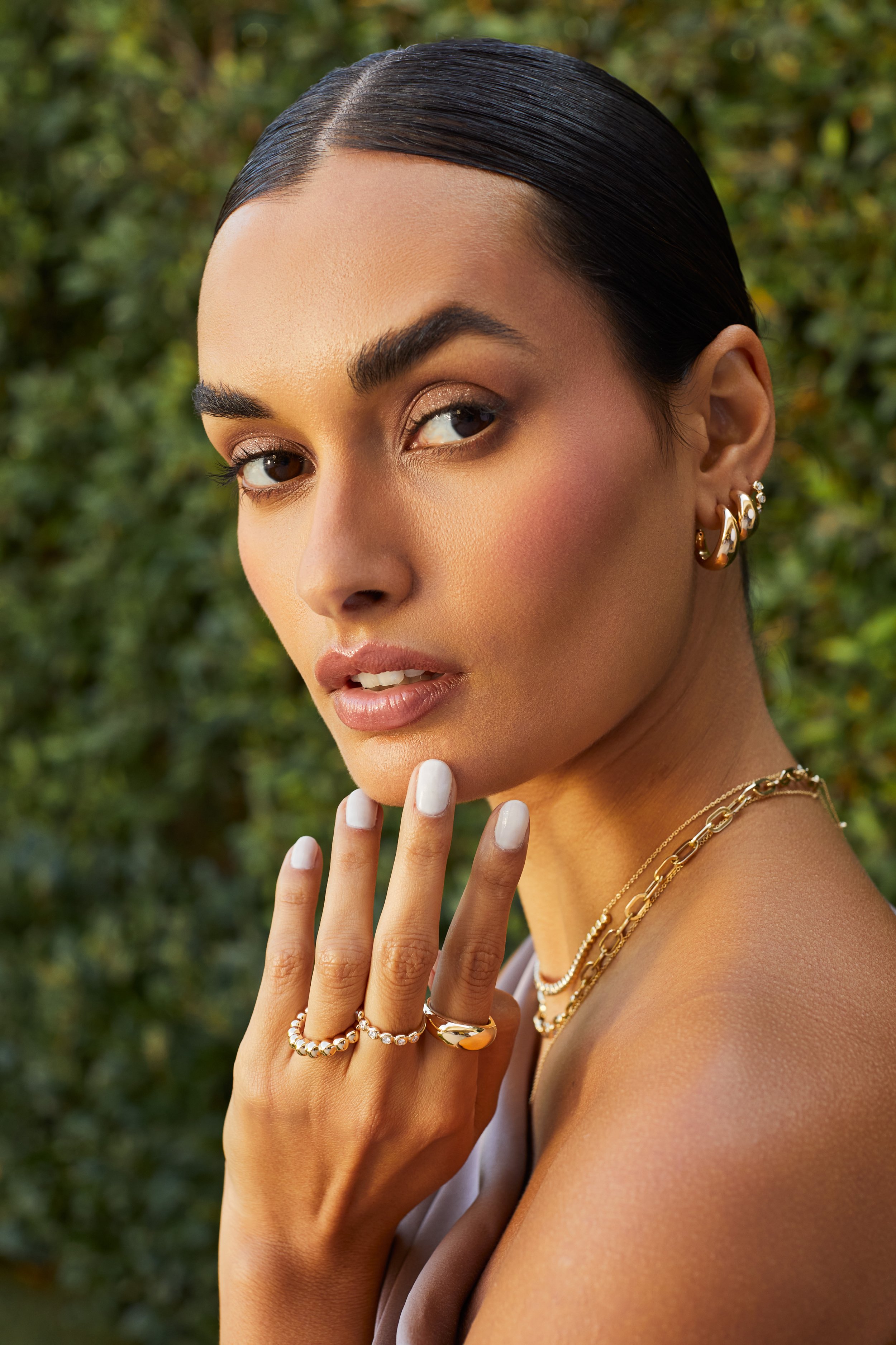 Close-up of a woman with dark hair slicked back, wearing gold earrings and layered gold necklaces, touching her face with manicured white nails, outdoors with blurred green foliage in the background.