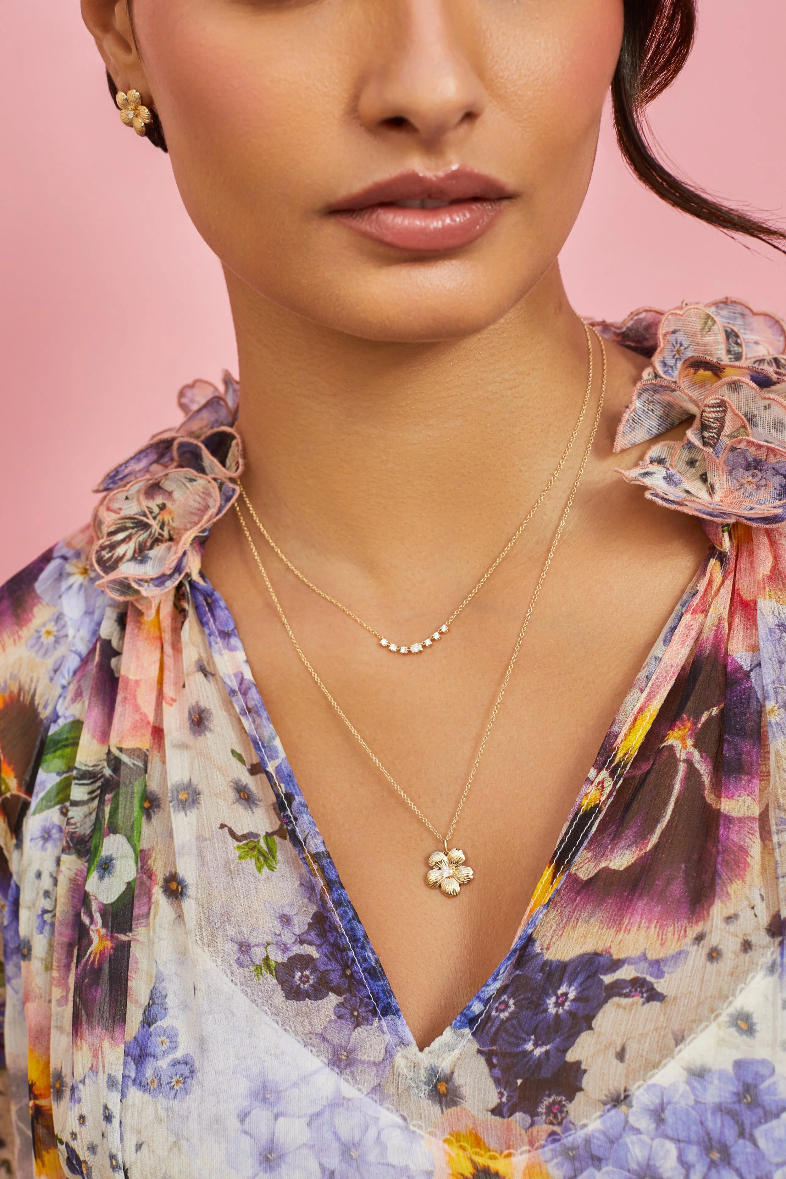 Close-up of a woman wearing layered gold necklaces with floral pendants, floral earrings, and a floral blouse with ruffled collar against a pink background.