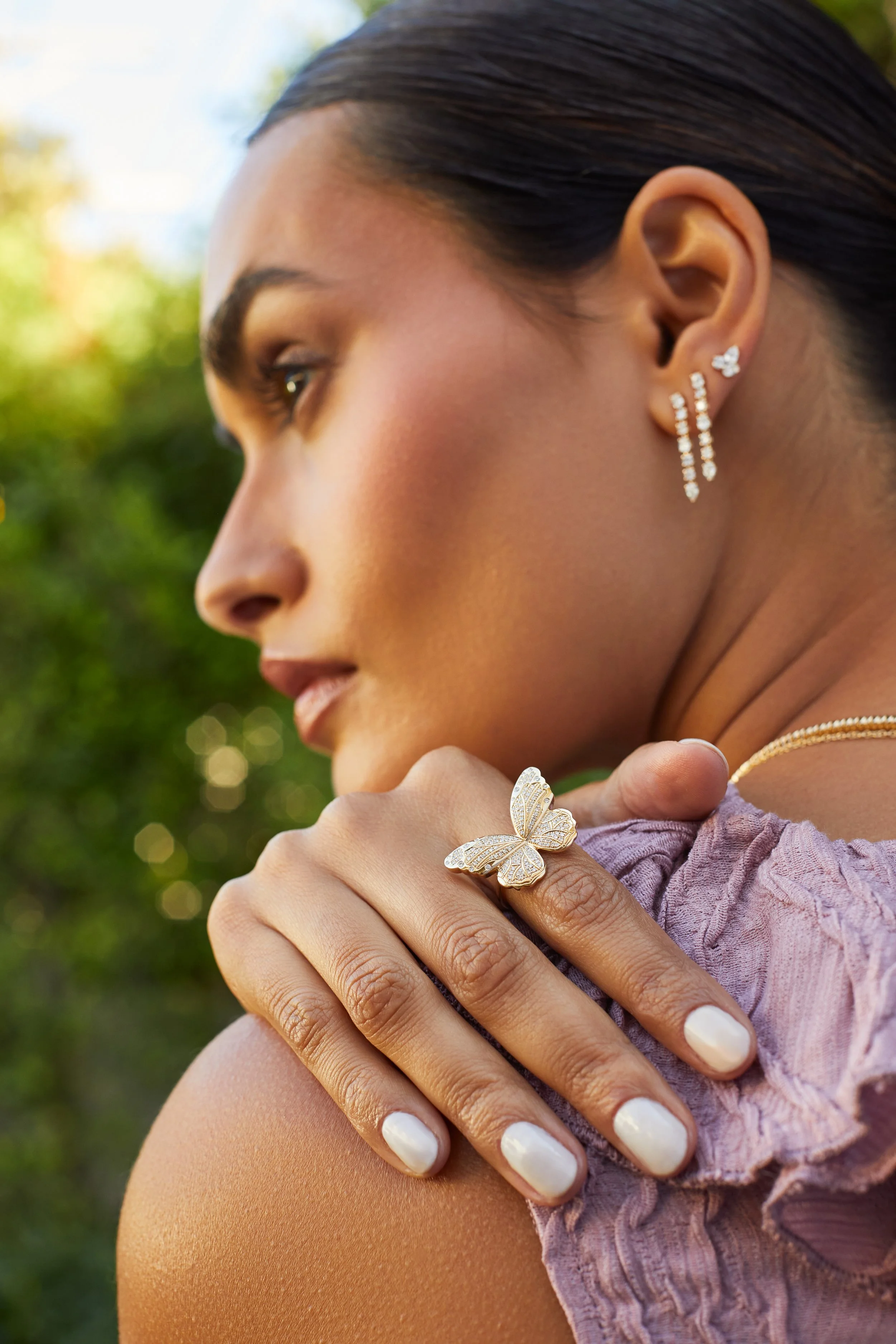 Close-up of a woman outdoors wearing jewelry, including butterfly-shaped rings, dangling earrings, and a necklace, with a blurred green background.