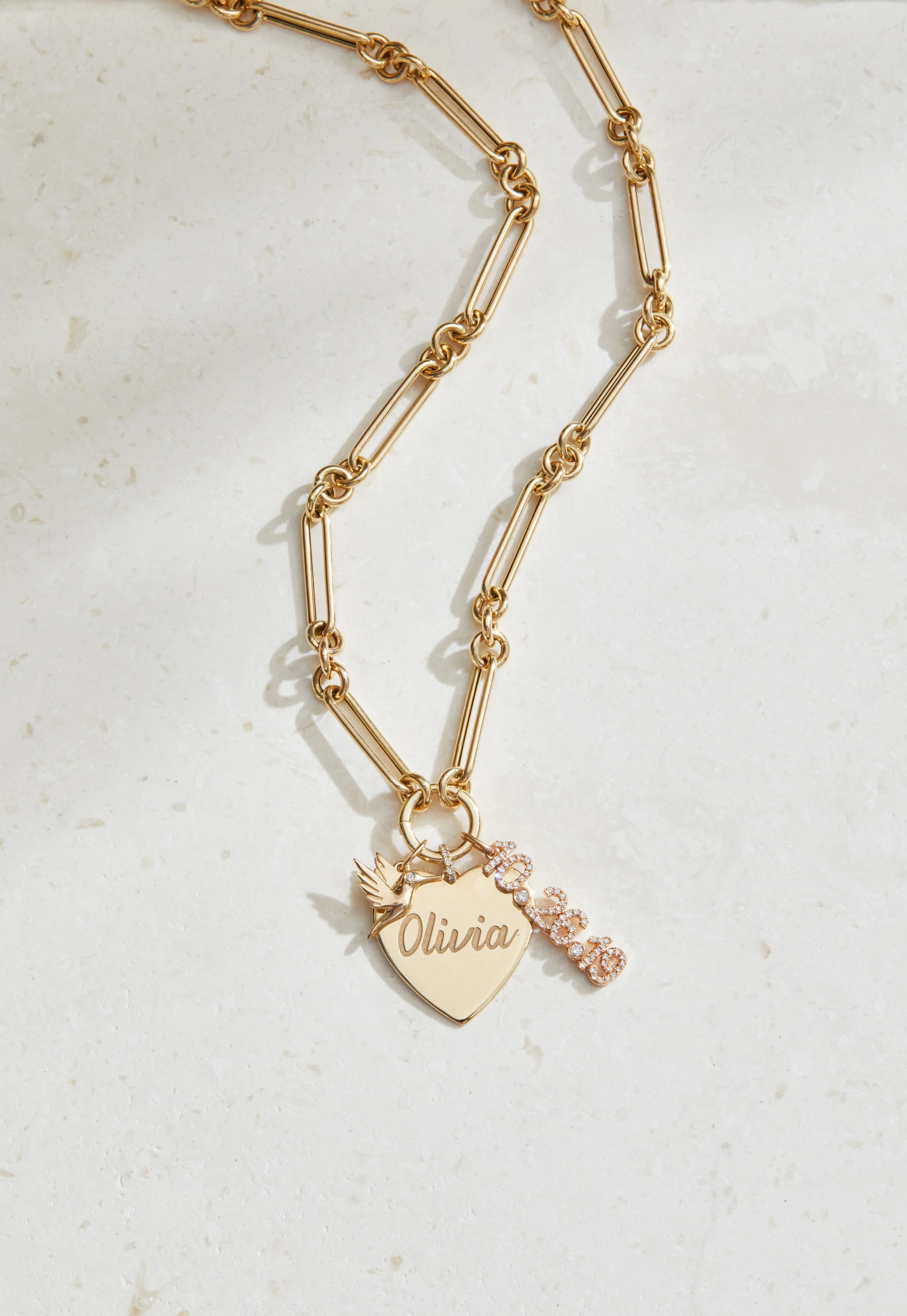 Gold chain necklace with two charms: a heart engraved with the name Olivia and a pink rhinestone-encrusted "love" charm, and a small bird charm.