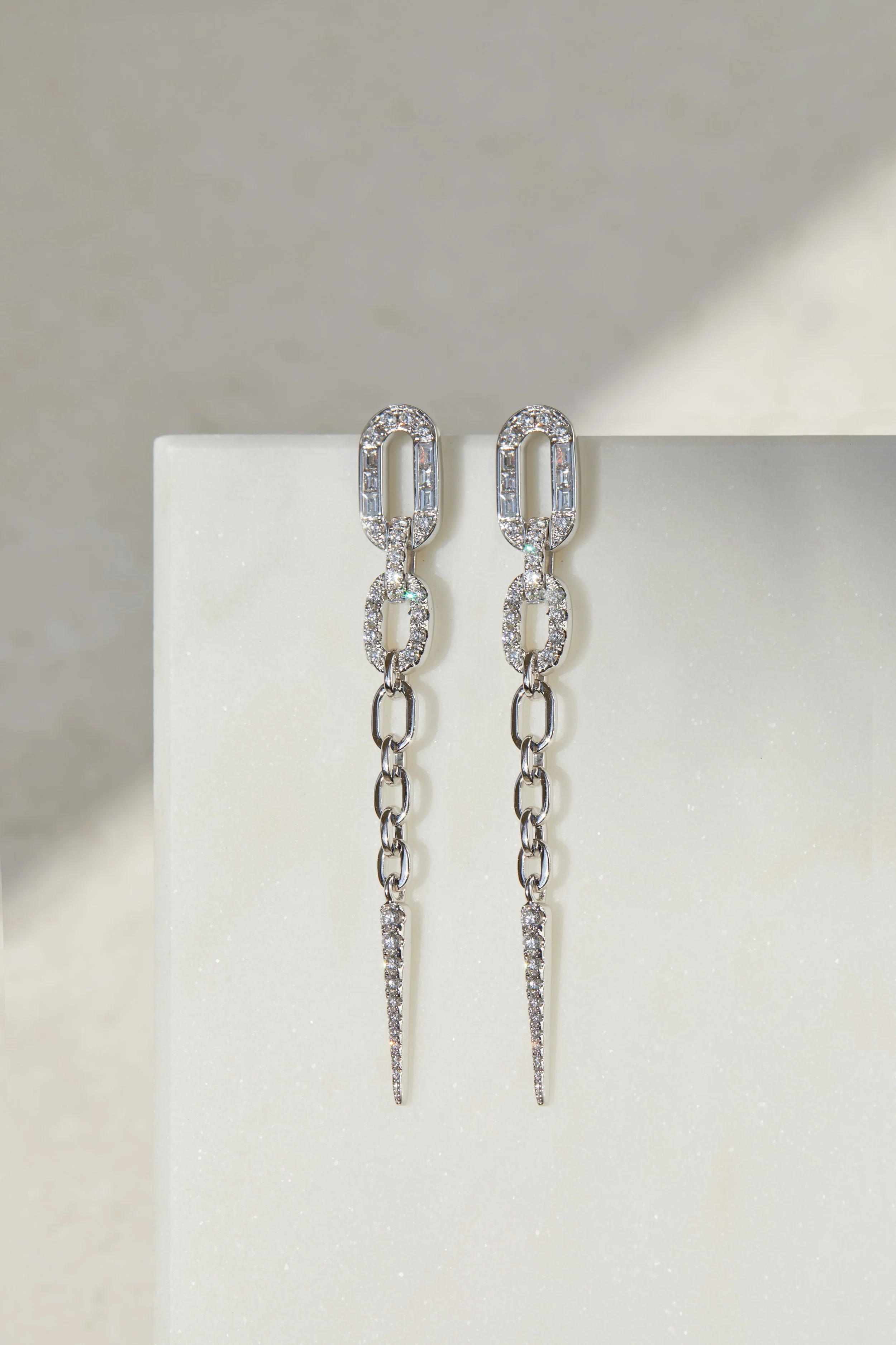Silver chain earrings with diamond accents hanging from a white surface.