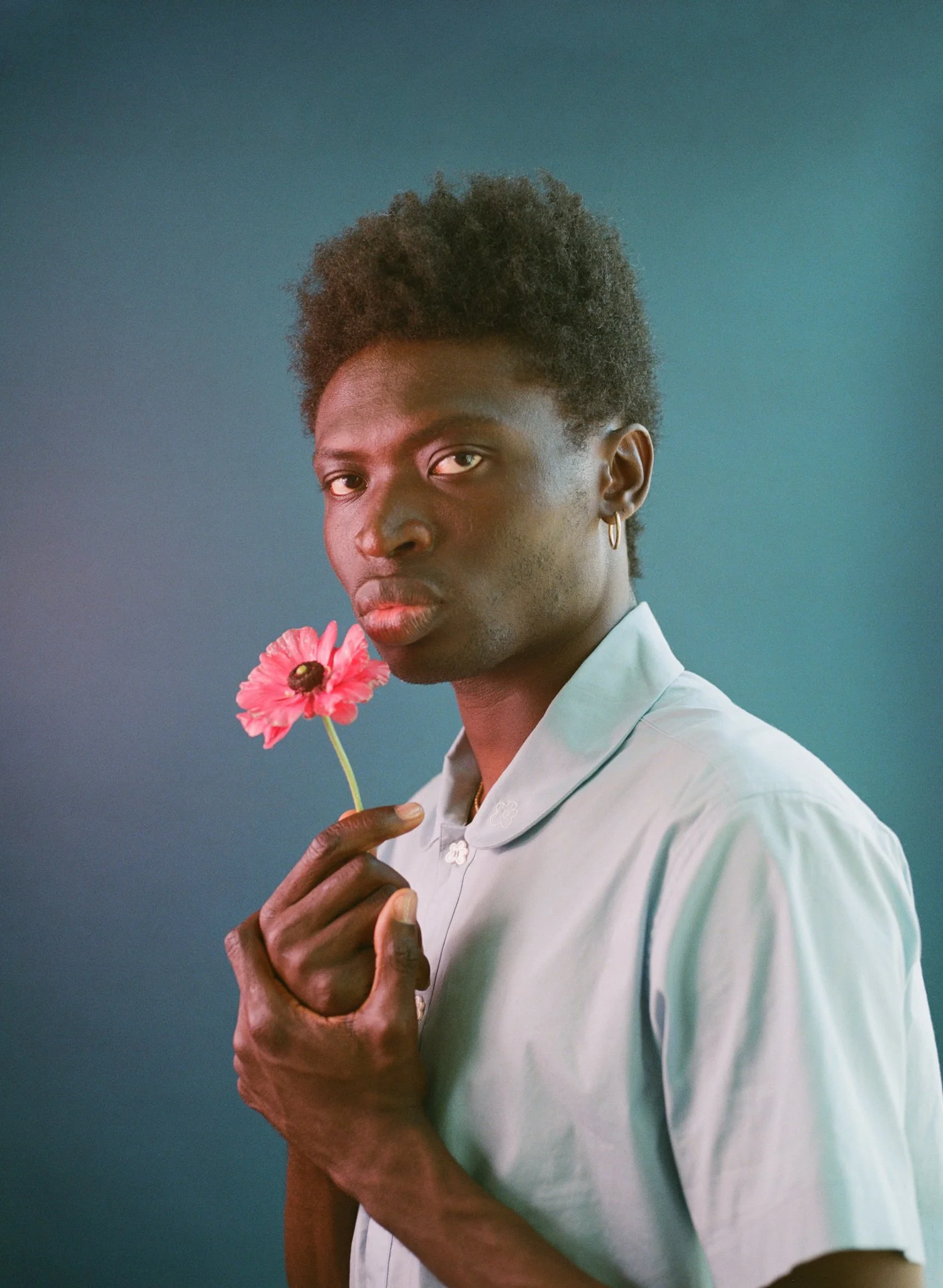 A young man with dark skin and short curly hair holding a pink flower near his face, wearing a light-colored shirt against a gradient background.