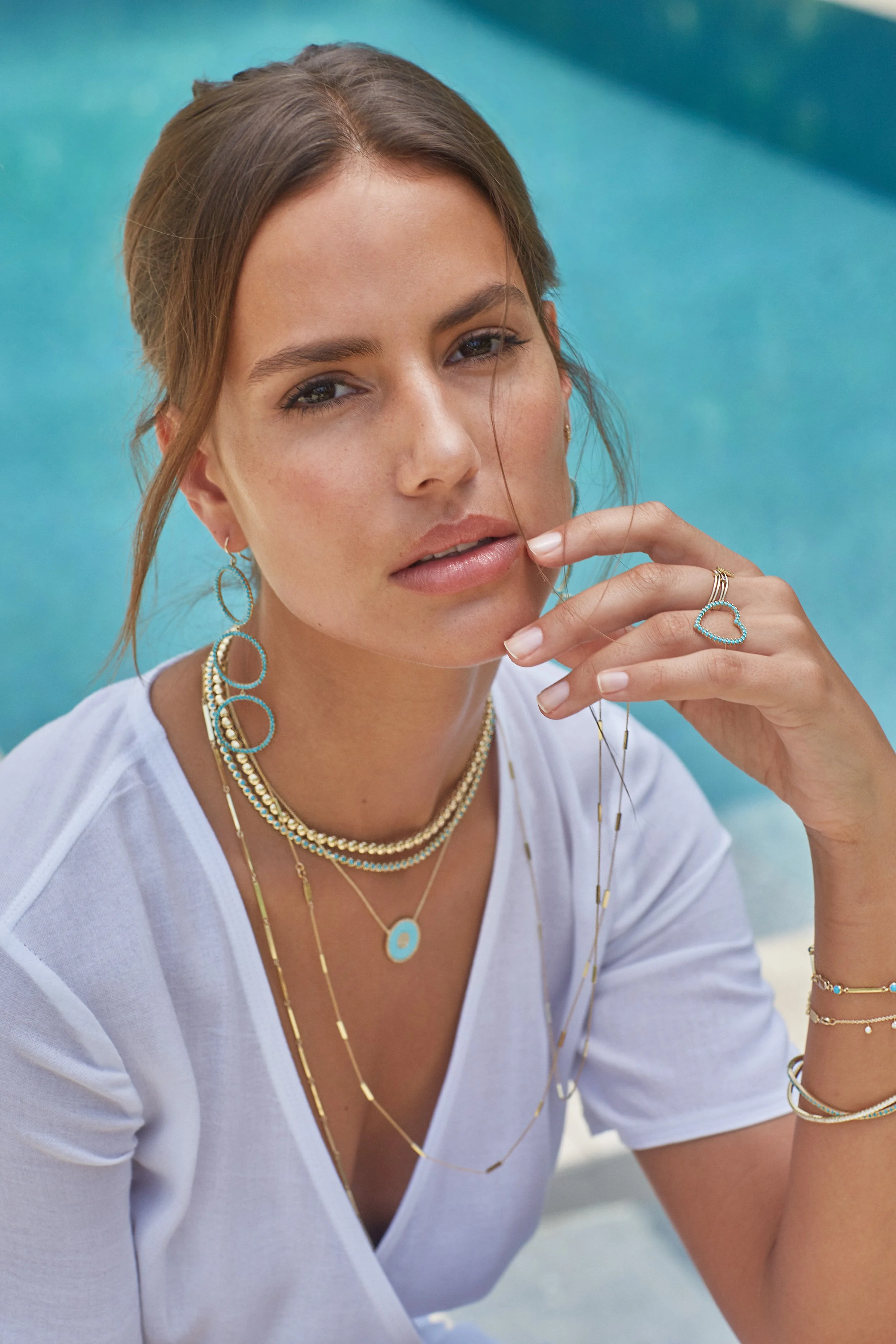 A woman with brown hair styled in a loose updo, wearing multiple layered gold and beaded necklaces, gold rings, and earrings, sitting by a pool with turquoise water in the background.