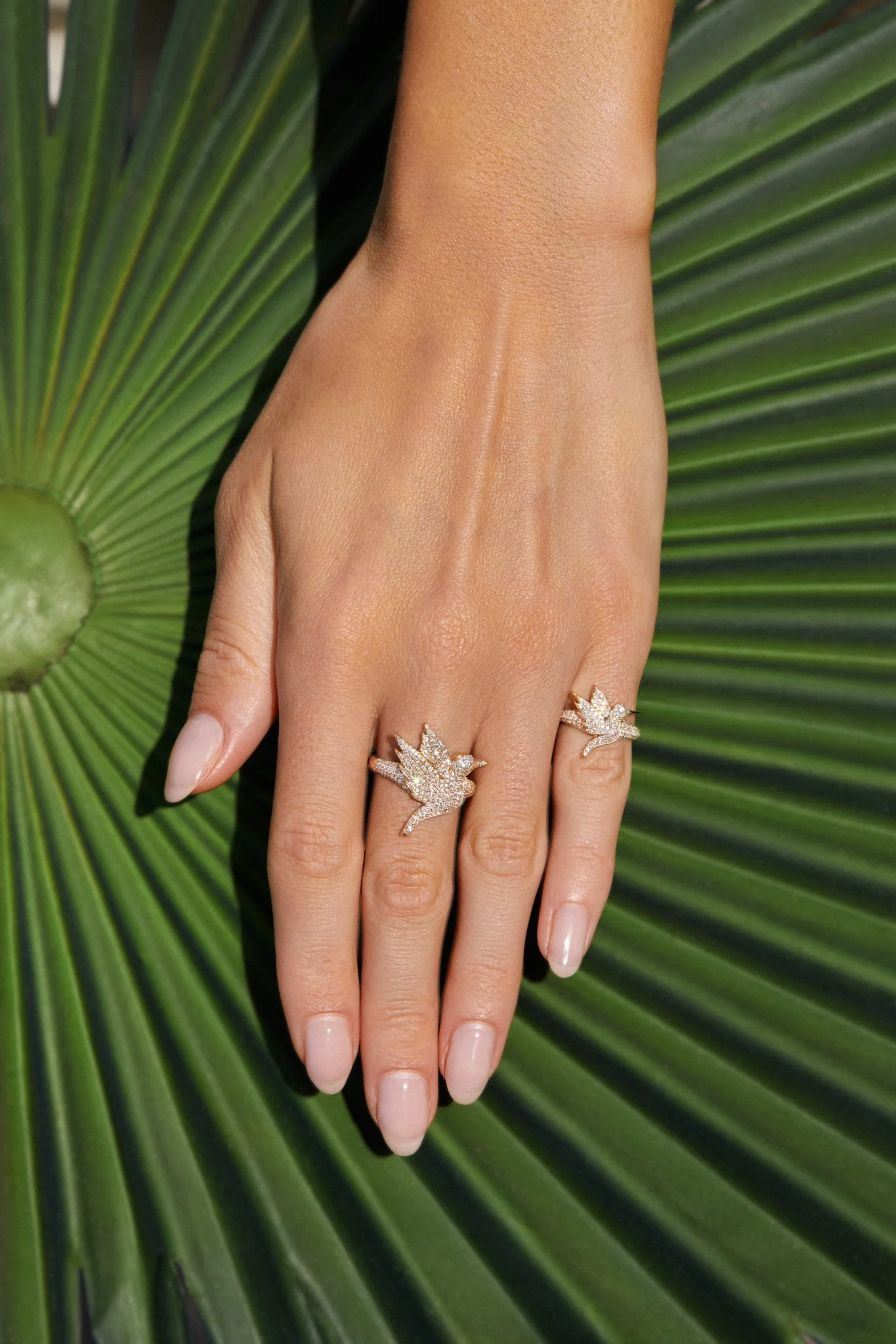 A hand with a manicured, natural-colored nail polish holding up two gold rings, each featuring a bird-shaped design adorned with diamonds, against a large green palm leaf background.