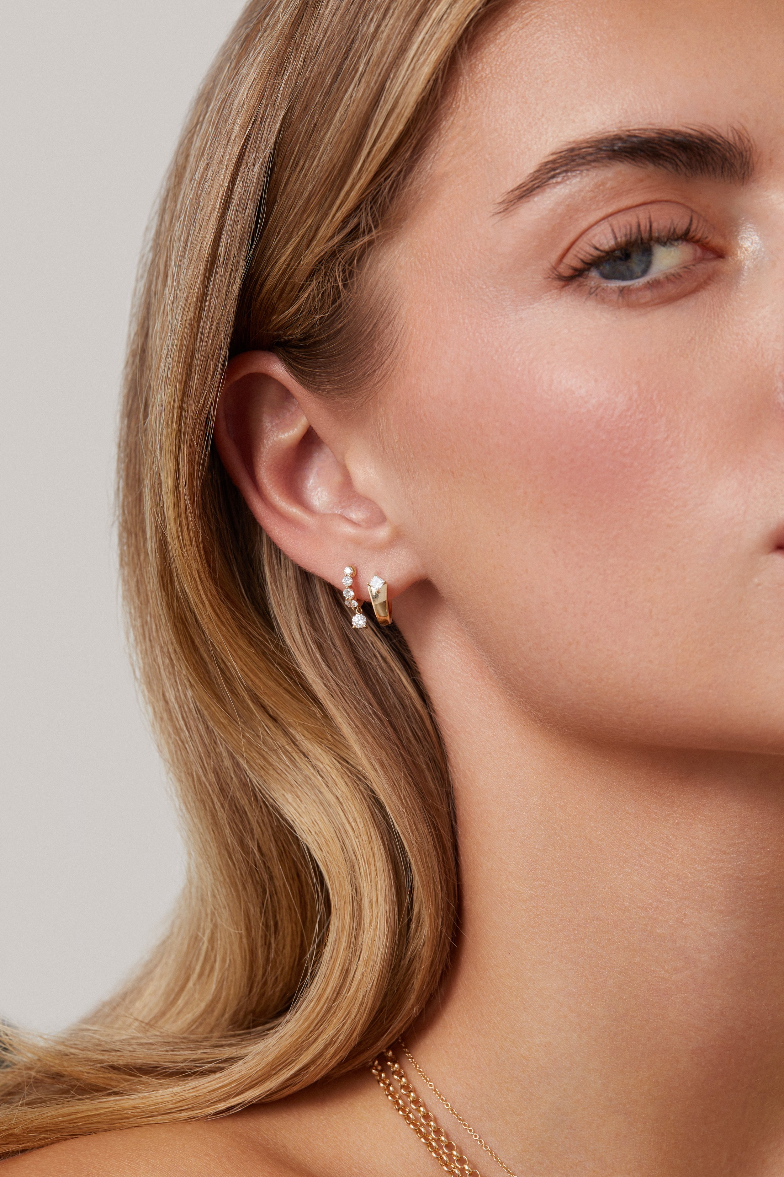 A close-up image of a woman's face showing her ear and part of her cheek, with blonde hair, wearing two gold hoop earrings with diamonds and a thin chain necklace.