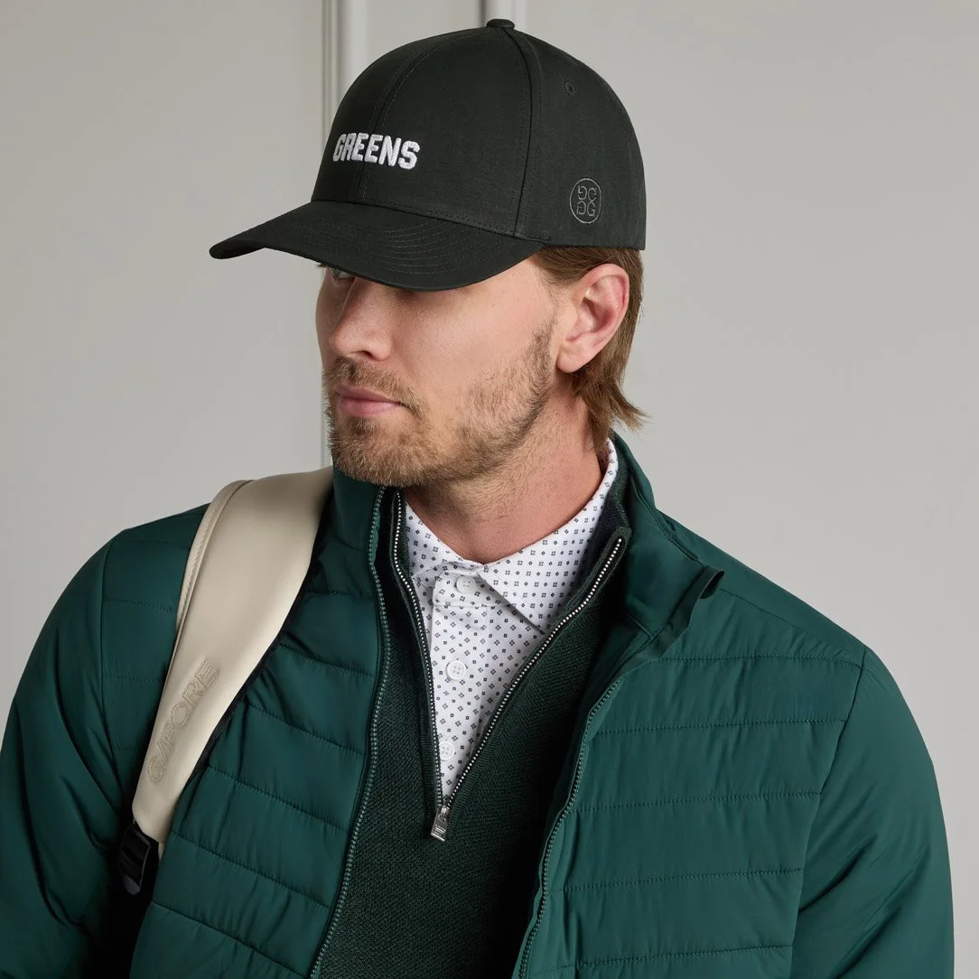 A man wearing a black baseball cap with 'GREENS' written on it, a green quilted jacket, a patterned white shirt, and a beige backpack.