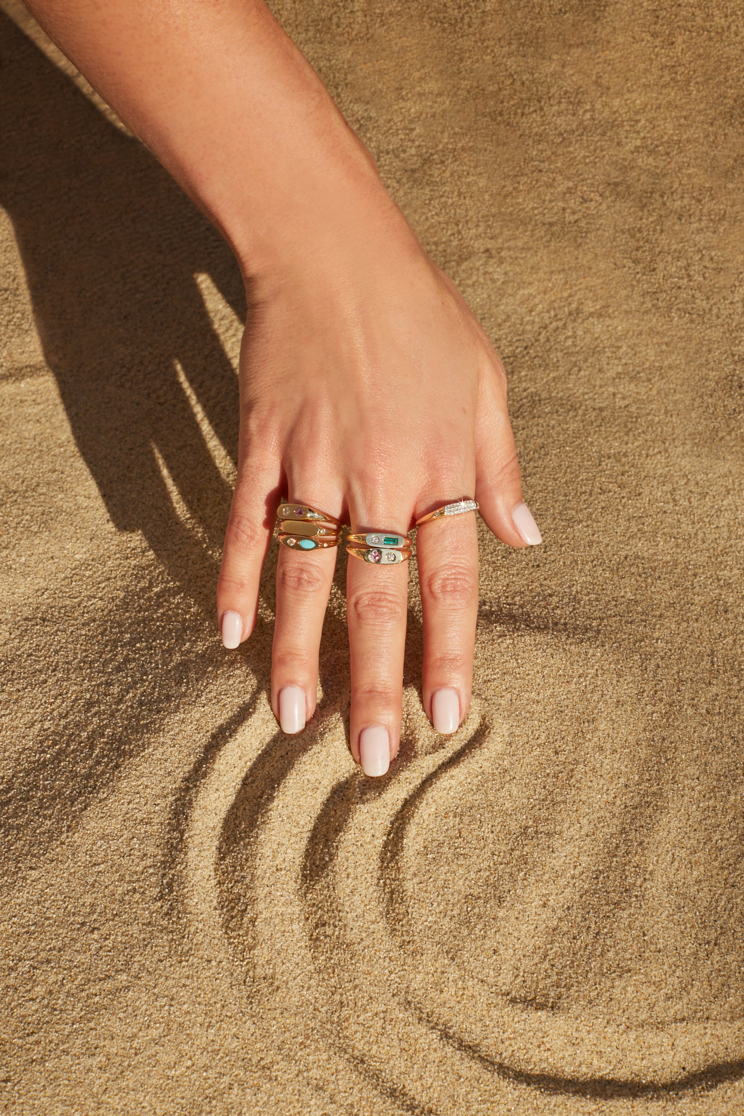 A close-up of a woman's hand with several rings on her fingers, resting on sand with ripples.