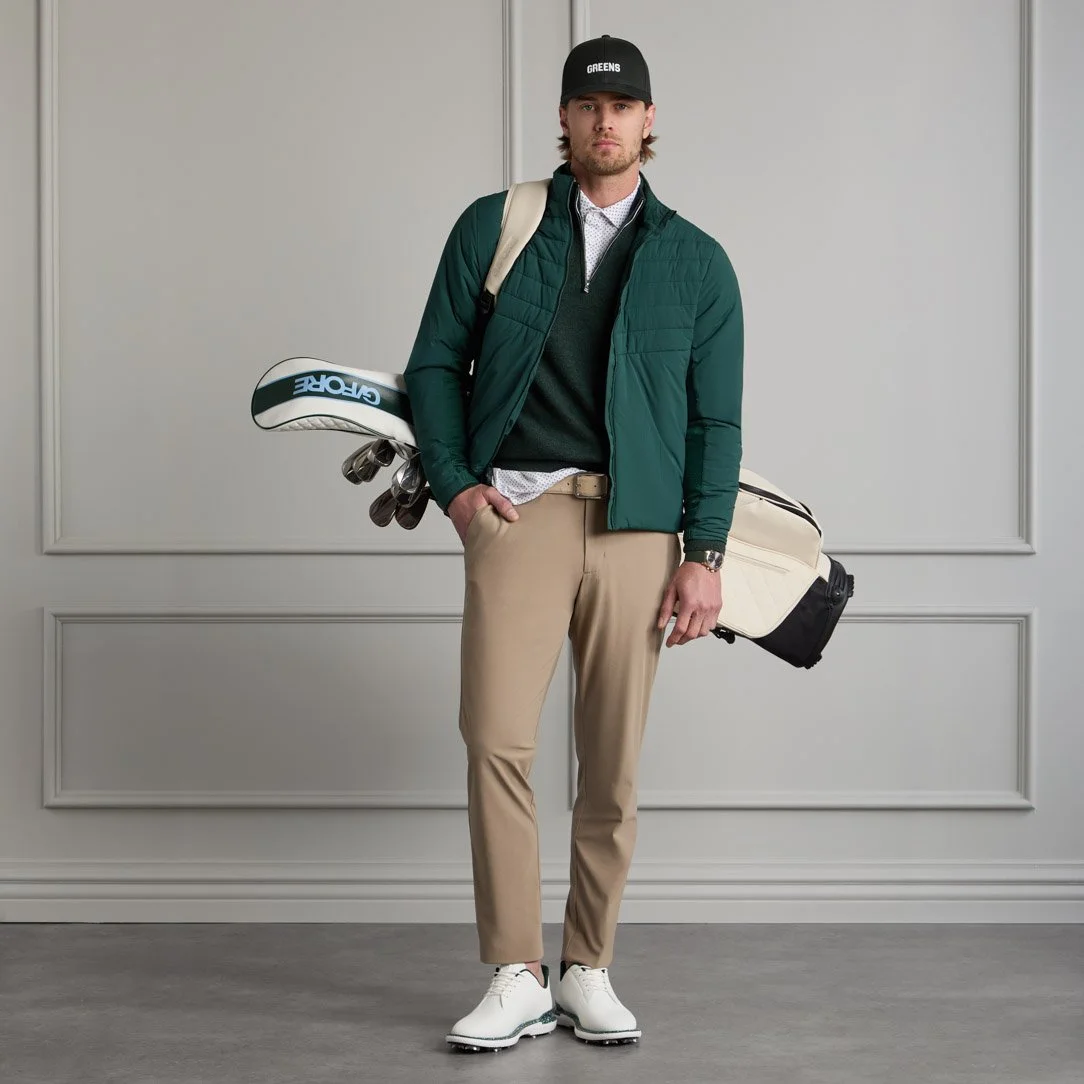 A man standing indoors, dressed in golf attire, holding a golf club on his left shoulder, wearing a black cap, green jacket, white shirt, beige pants, white shoes, and carrying a golf bag.