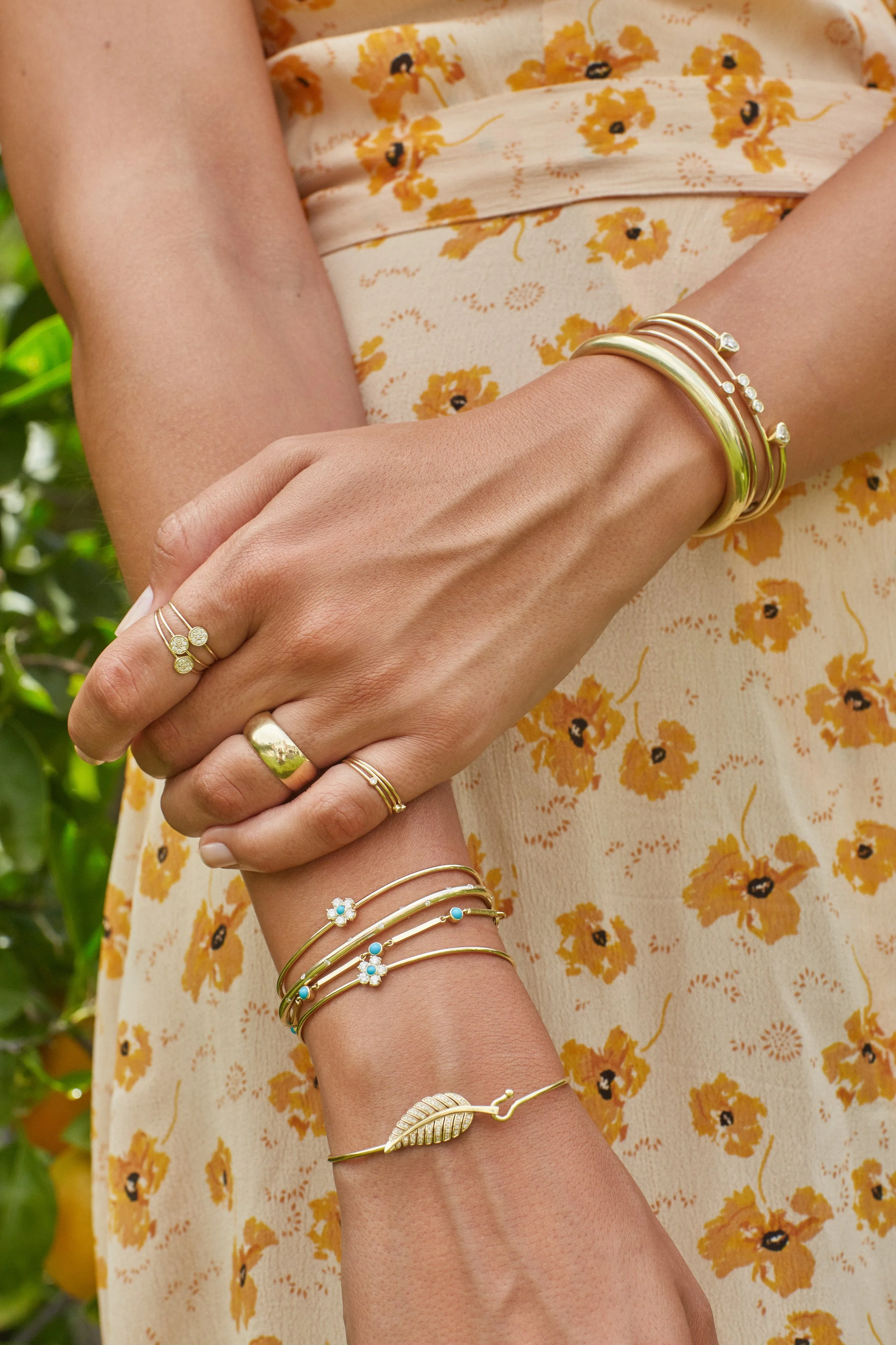 A woman is wearing multiple gold jewelry bracelets and rings on her hands and wrists, with her arms crossed. She is dressed in a light-colored dress with a yellow floral pattern, and the background appears to be green foliage.