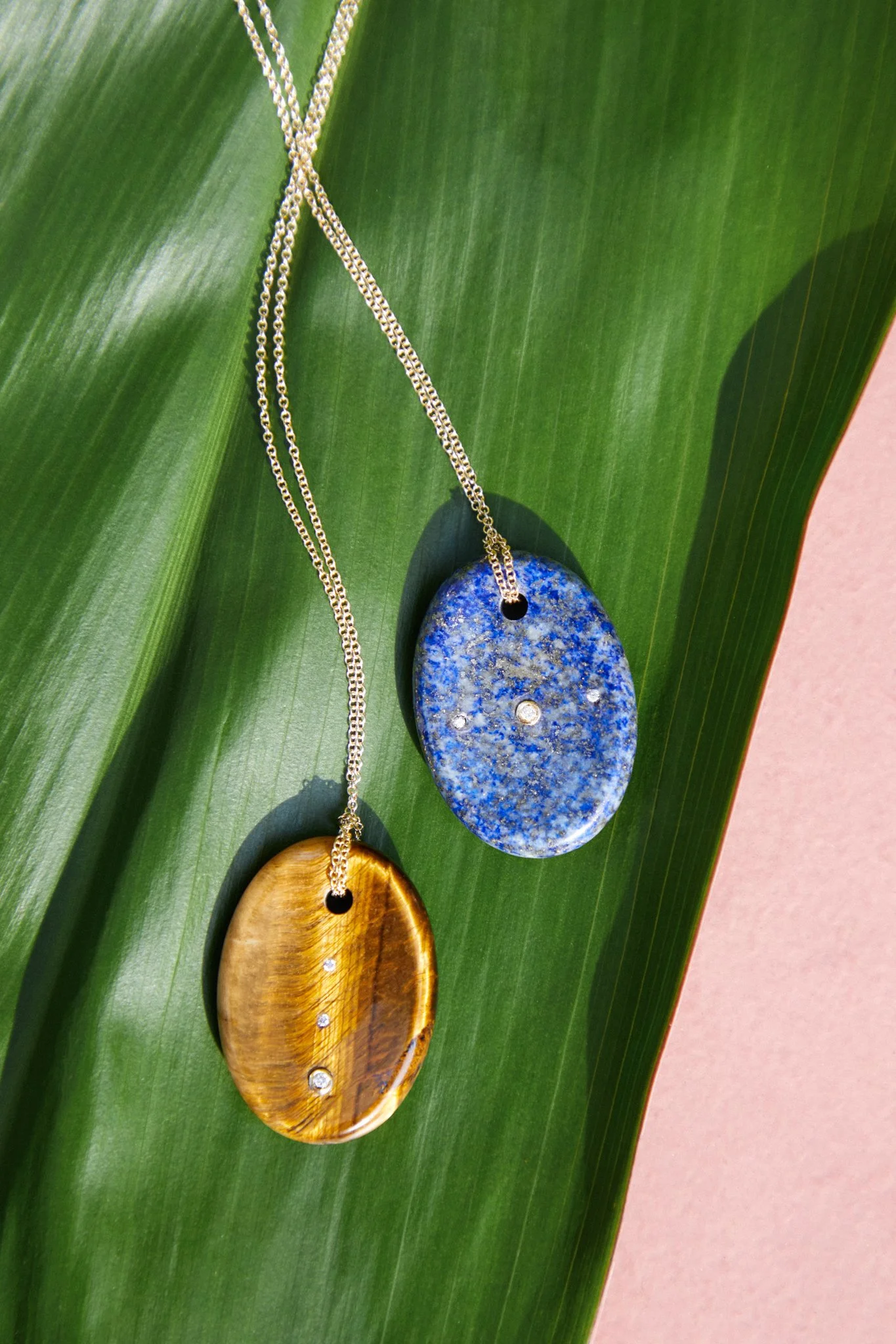 Two pendants, one blue and one golden, with small embedded stones, hanging on silver chains over a green leaf.