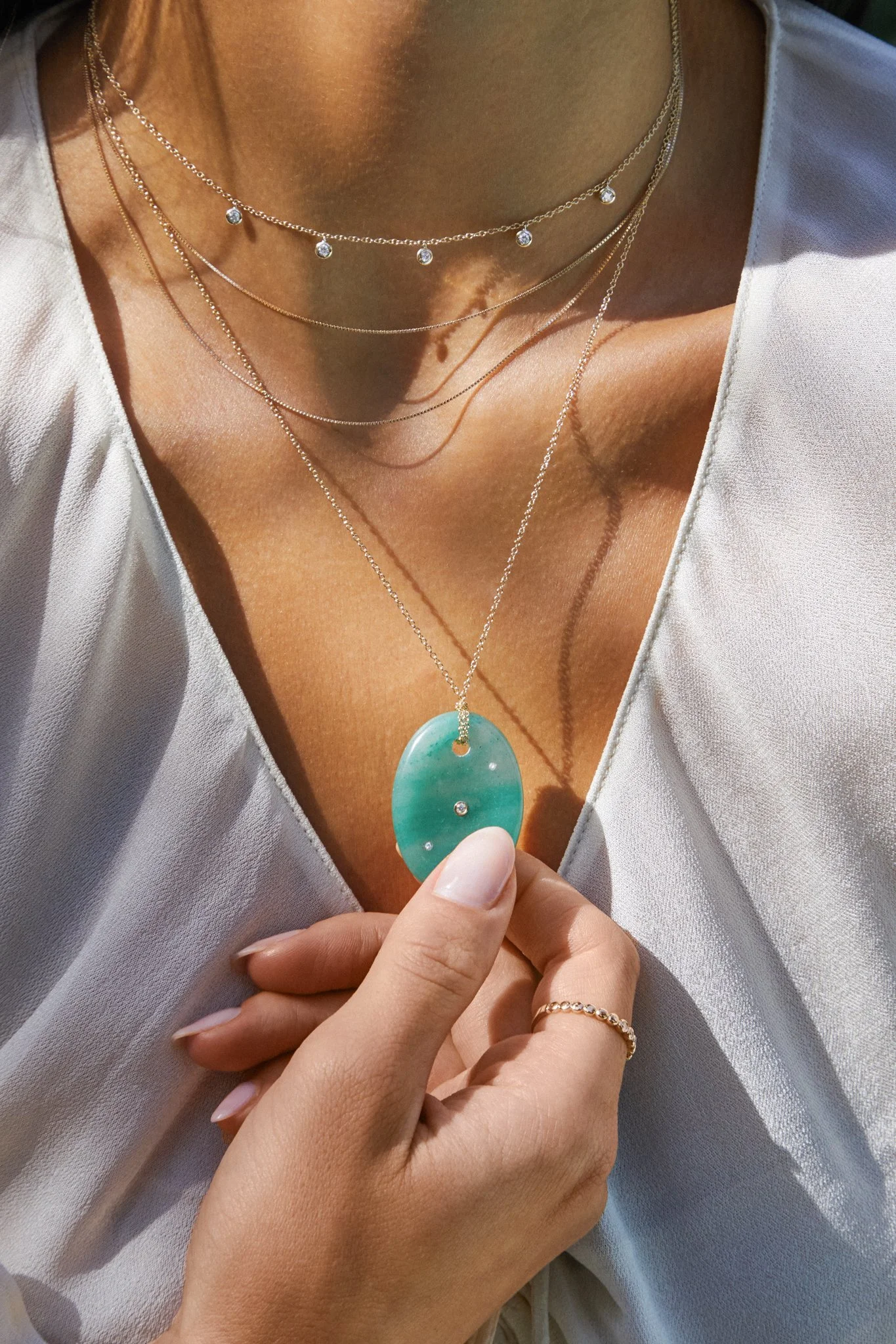A woman holds a turquoise pendant necklace with a white dress and layered gold chains.