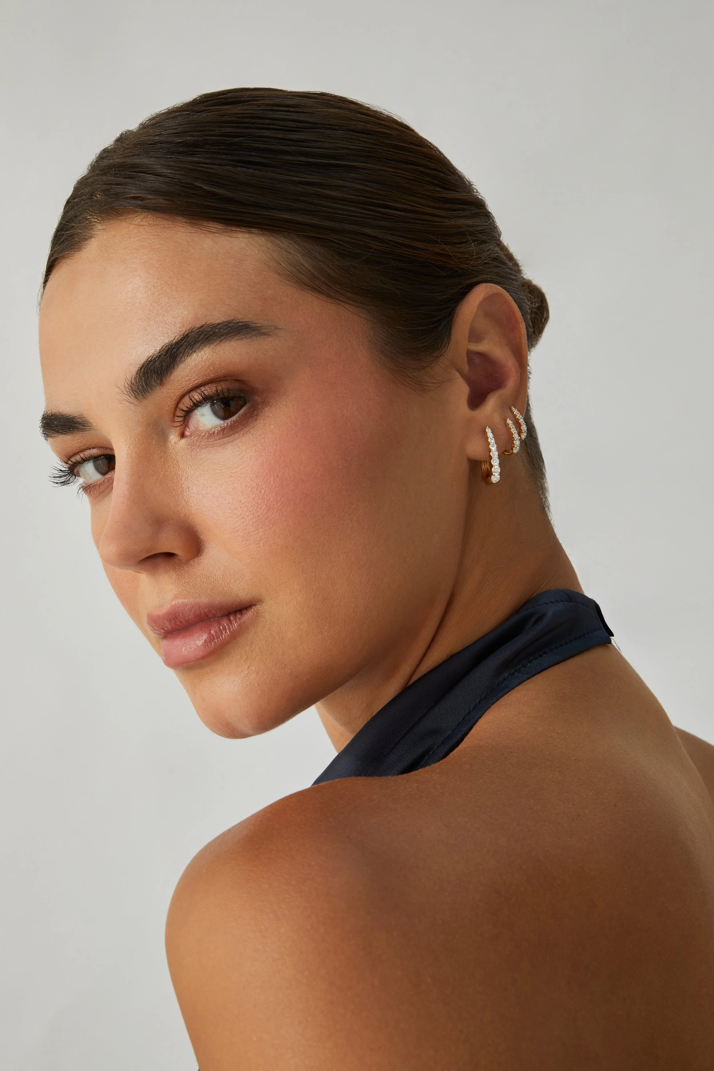 A woman with brown hair slicked back, wearing three diamond earrings in her right ear, shown in close-up against a plain white background.