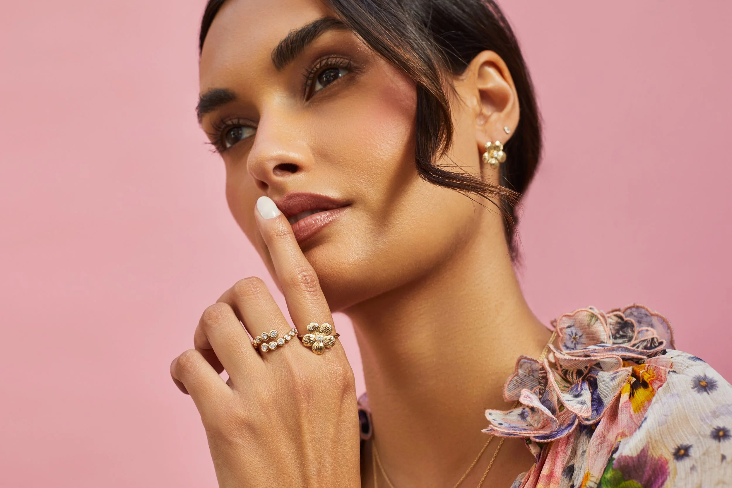 A woman with short dark hair and makeup, wearing floral clothing and gold jewelry, posing with her index finger near her lips against a pink background.
