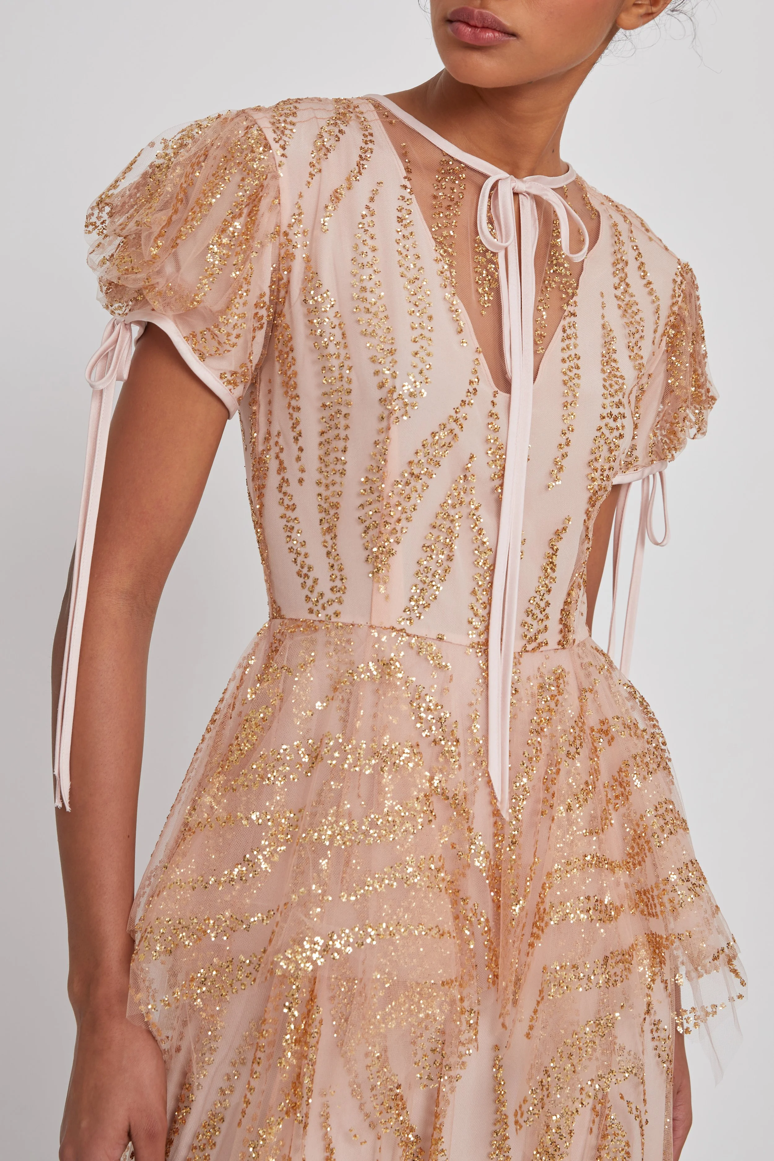 A woman wearing a pink dress with gold embroidery and tulle fabric, featuring puffed short sleeves with tied ribbons and a ribbon tie at the neckline.