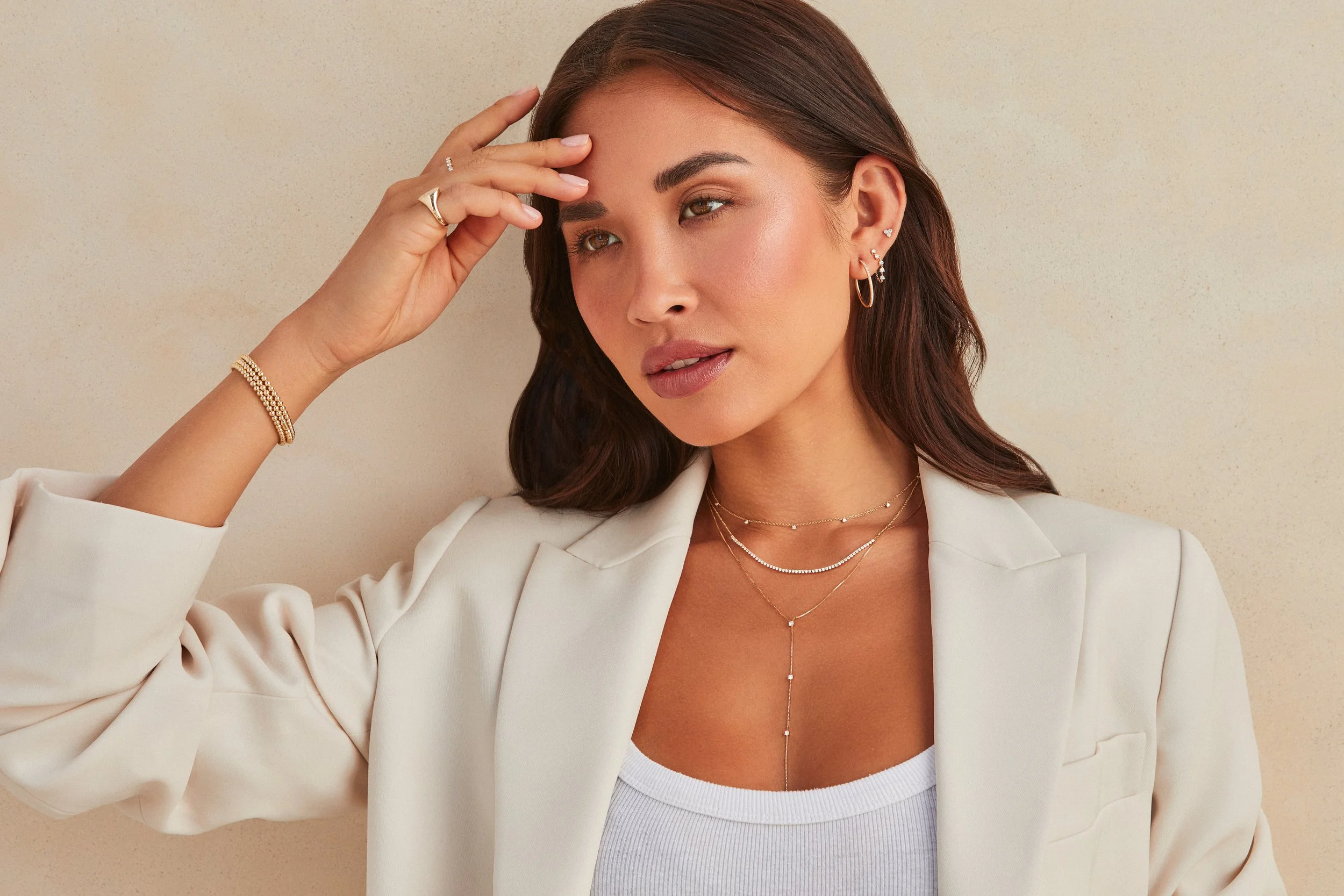 A woman with brown hair and light makeup wearing a white blazer and layered jewelry, posing with her hand on her forehead against a beige background.