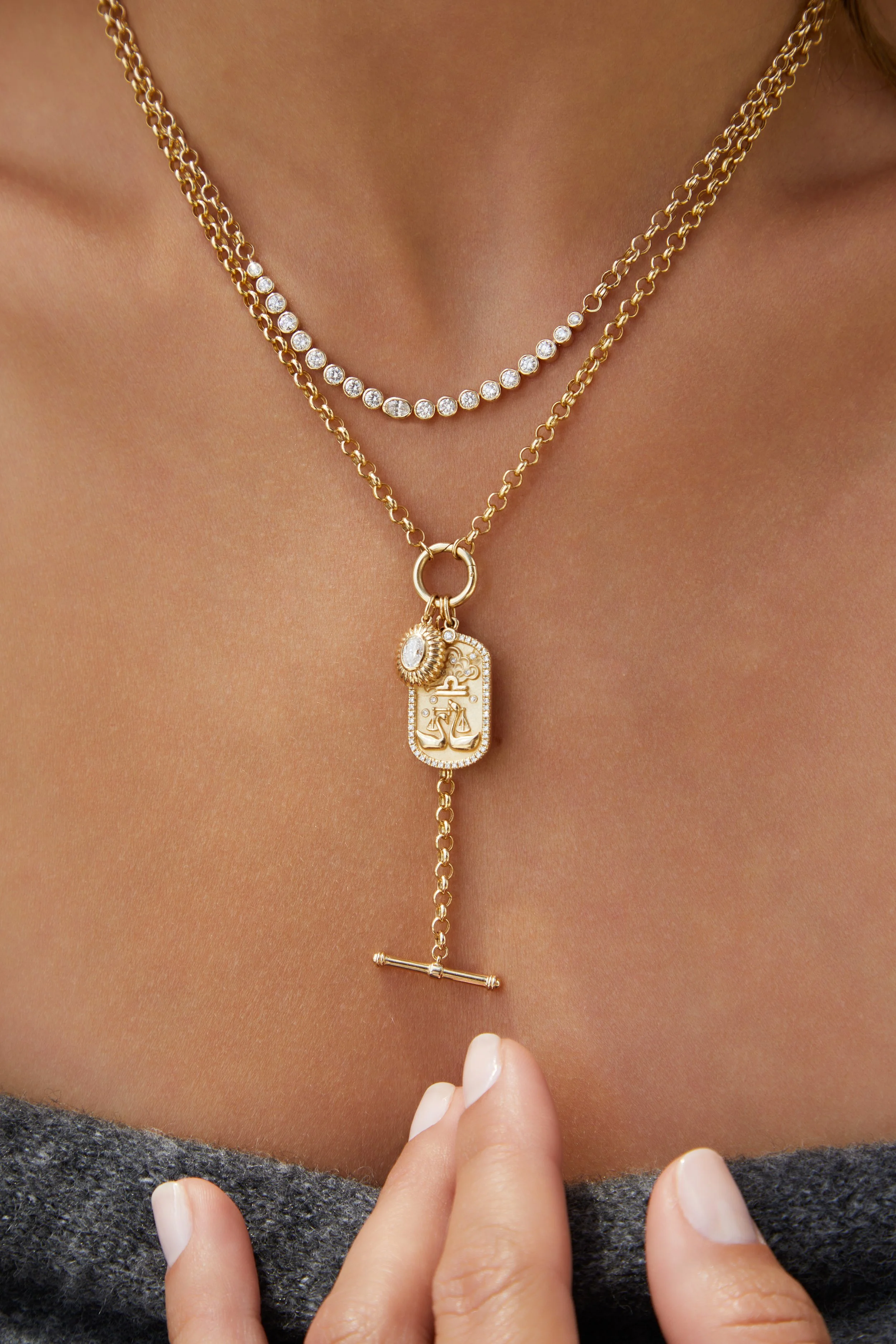Close-up of layered gold necklaces on a woman's neck, featuring a pendant with a scale and anchor emblem, a gemstone, and a bar charm, with a person touching the lower necklace.