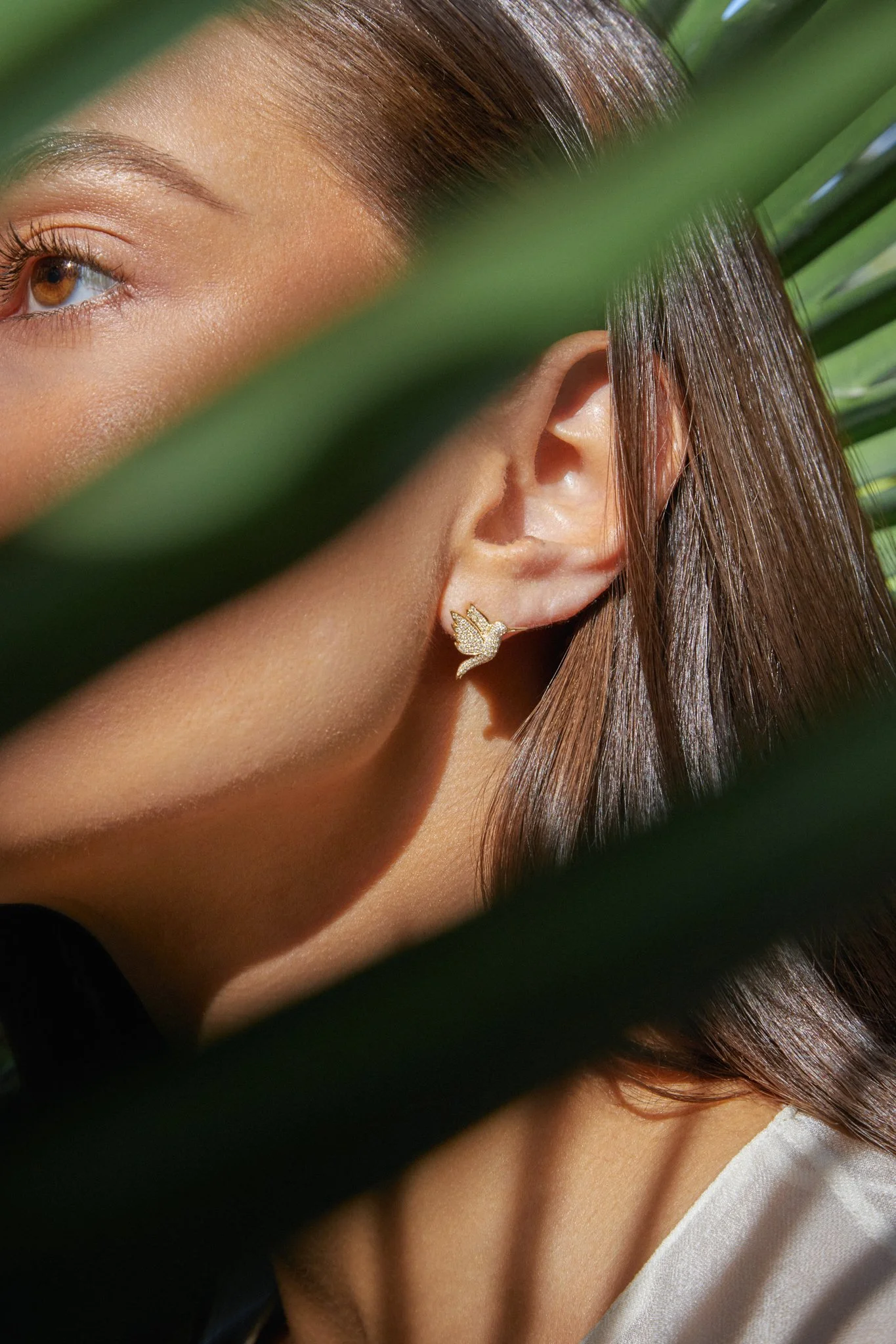Close-up of a woman's face and ear, showcasing a gold swan-shaped earring, partially obscured by green leaves with sunlight highlighting her skin and hair.
