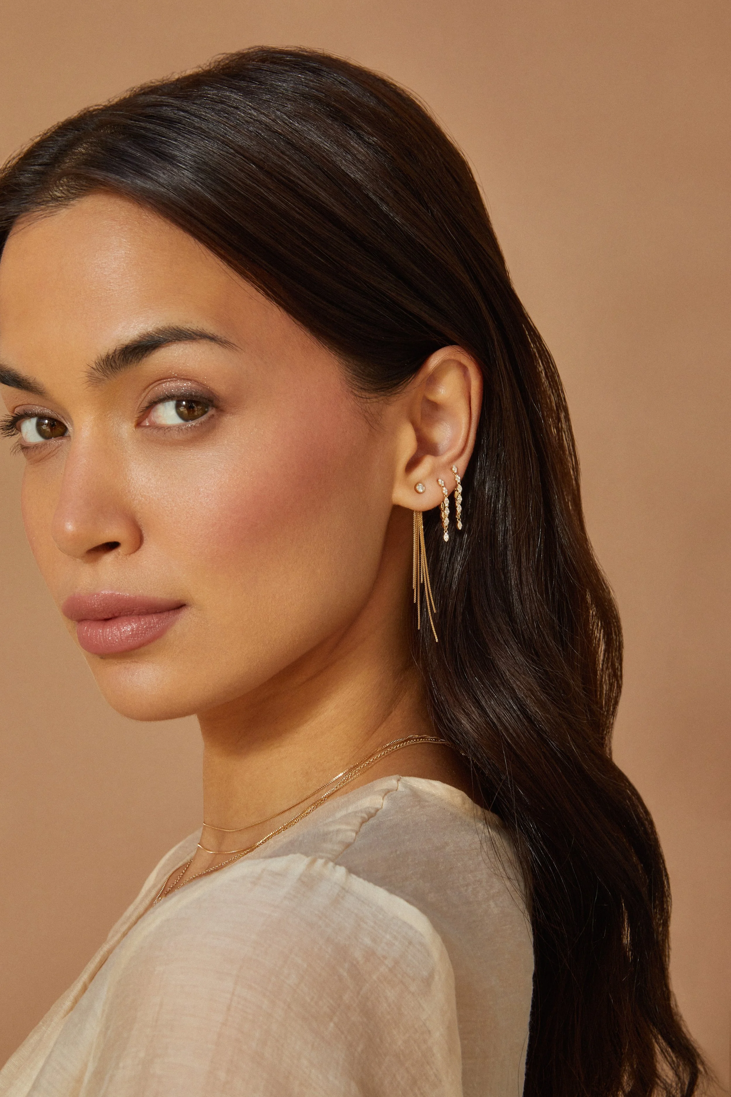 Close-up of a woman with smooth, dark hair and light makeup, wearing multiple earrings and gold necklaces, against a beige background.