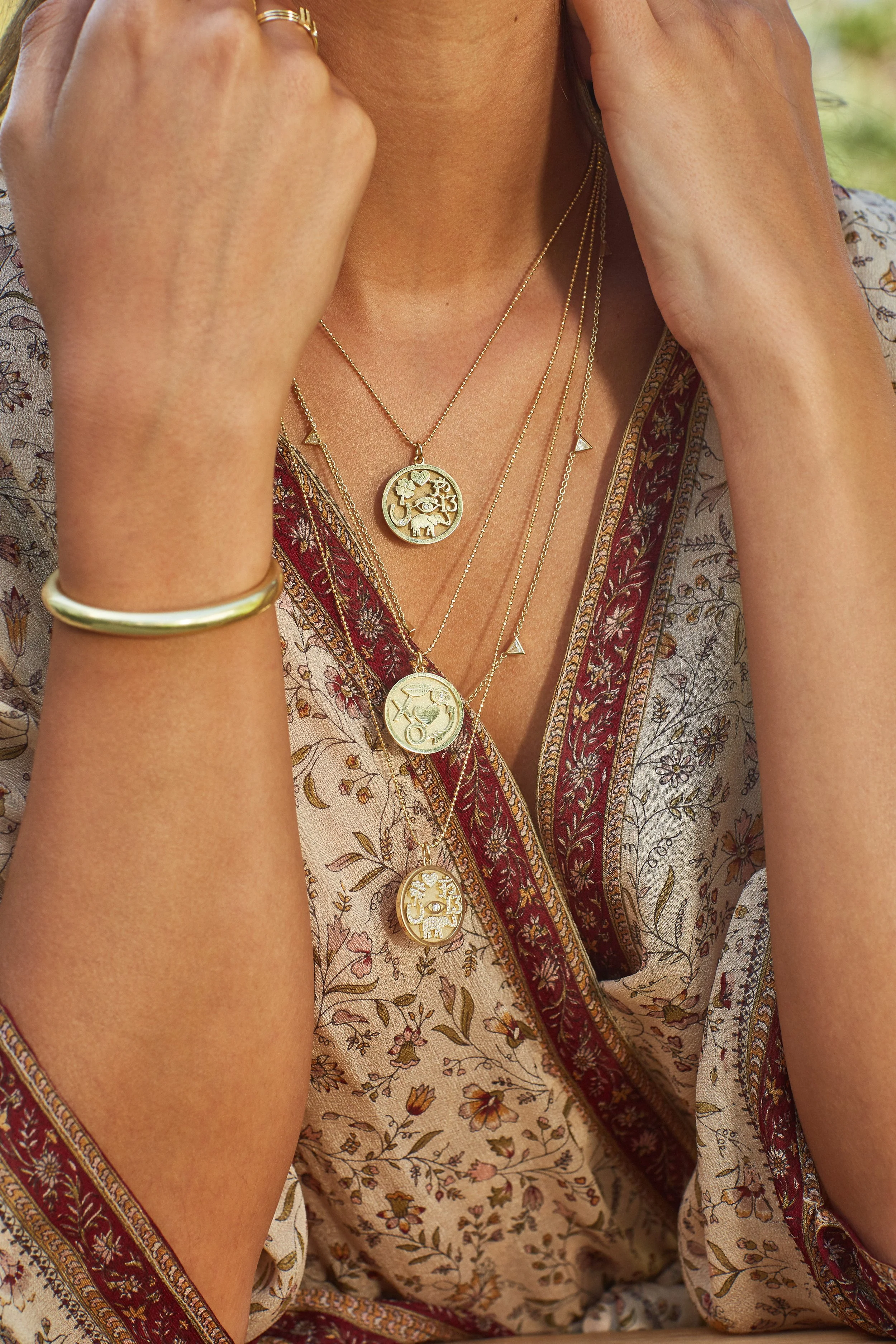 Multiple layered gold necklaces with circular pendants, gold bracelet on left wrist, woman wearing floral patterned dress with red and beige borders.