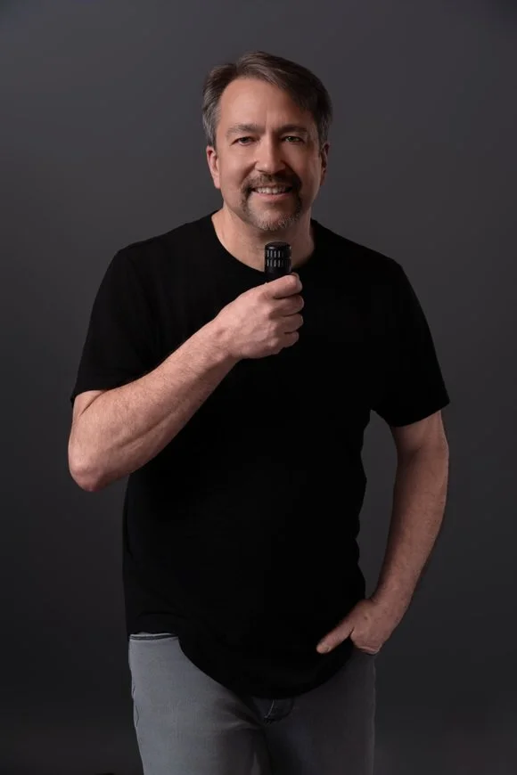 Headshot of a man in a black shirt holding a microphone