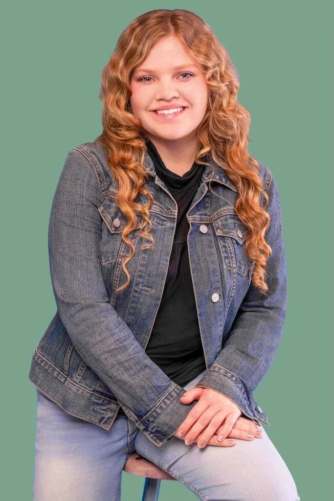 A teenage girl with long, curly red hair wearing a denim jacket with a black shirt underneath and jeans. She is smiling and posed with her hands on her knee, partially seated on a stool.