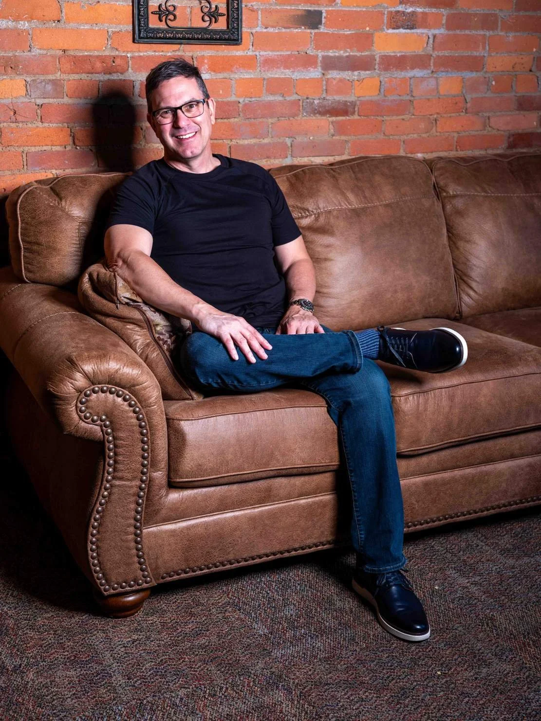 Headshot of a white middle aged man with glasses wearing a plain black shirt and blue jeans seated on a brown leather couch