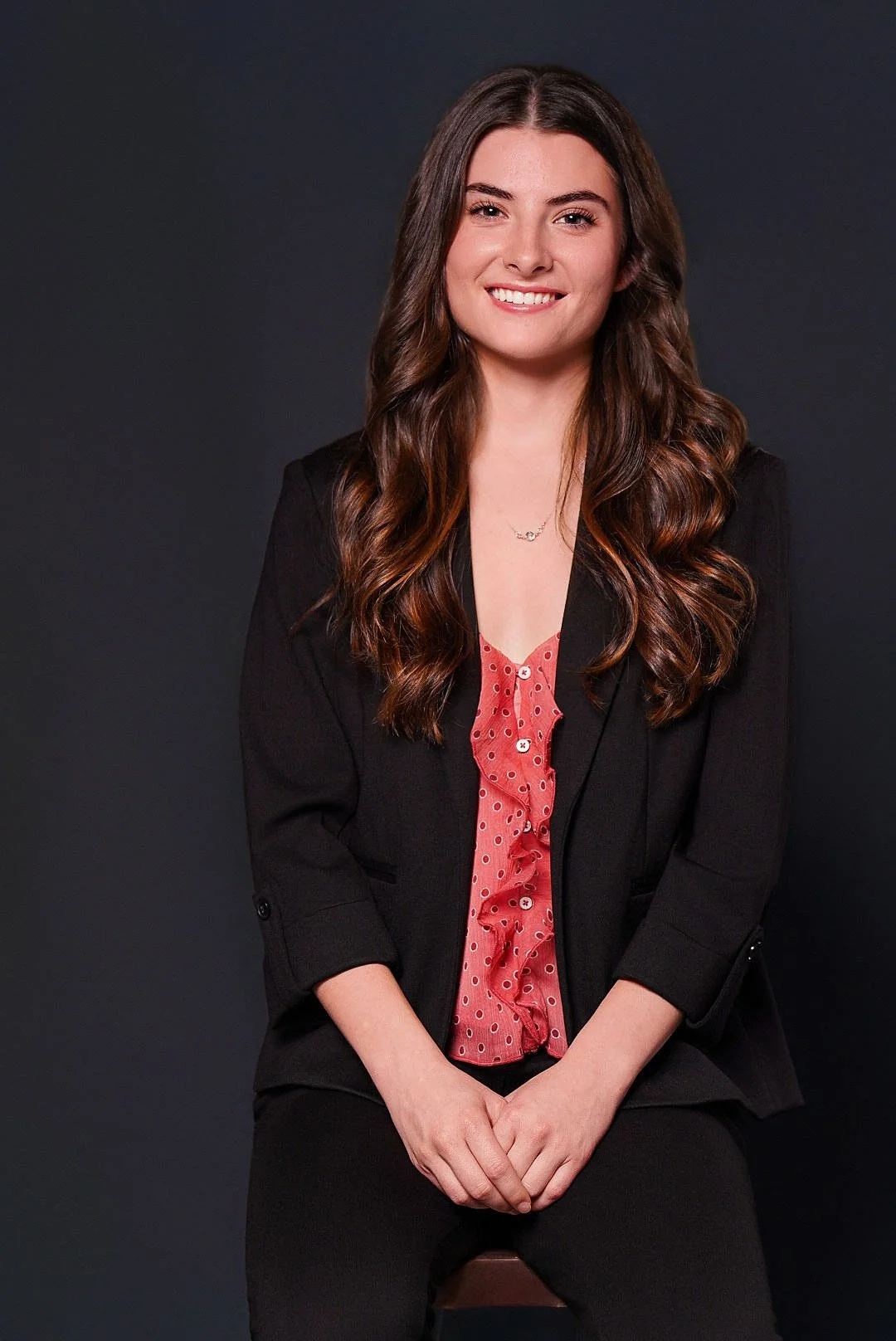 A young woman with long brown hair seated with her hands in her lap. She is smiling and wearing a black blazer with a pink undershirt and black pants
