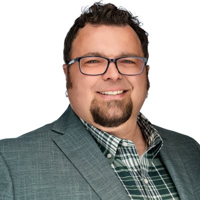 Headshot of a man wearing a grey jacket with a plaid button up underneath, glasses, and a smile