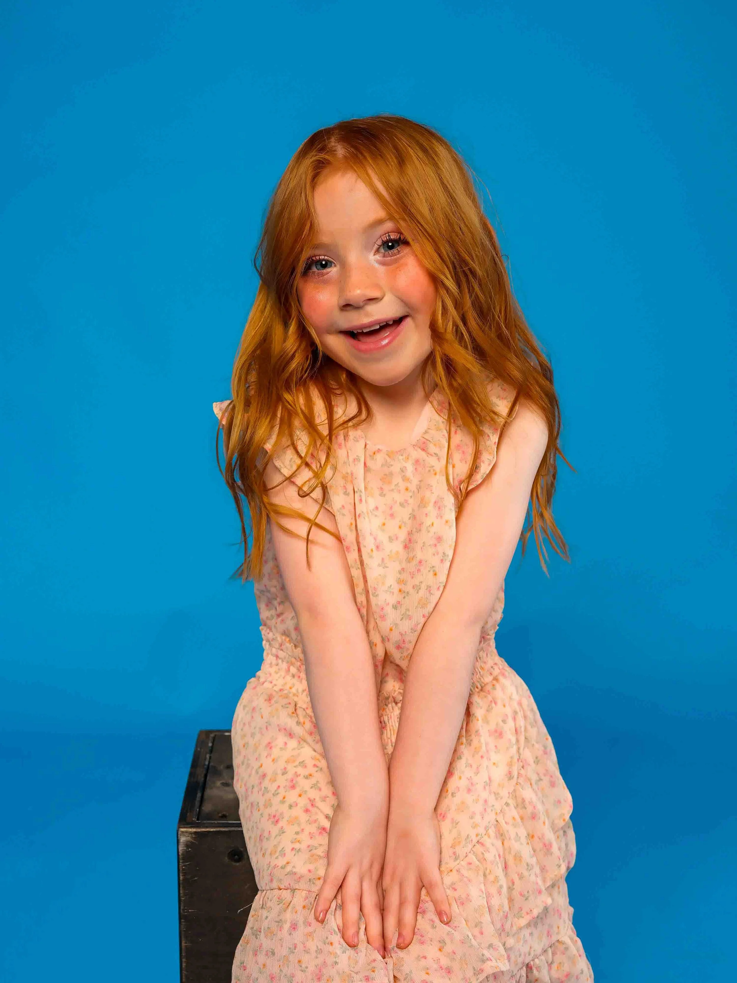 A young girl with orange hair, rosy cheeks, and a pink floral dress seated on a wooden box. She has her hands in her lap and is smiling/