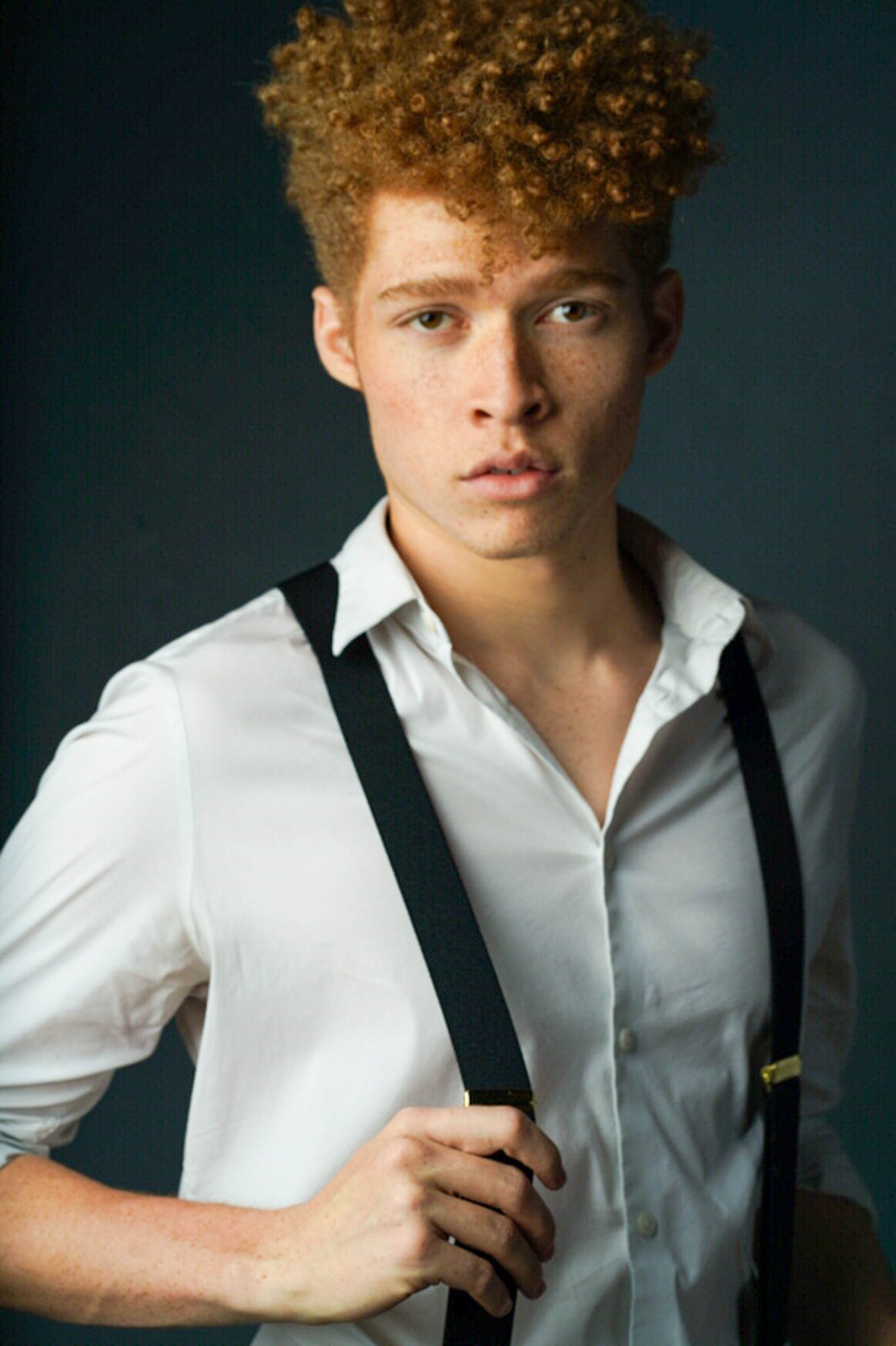 Headshot of a man with short, curly, orange hair wearing a white button up shirt with black suspenders, one of which he is holding.