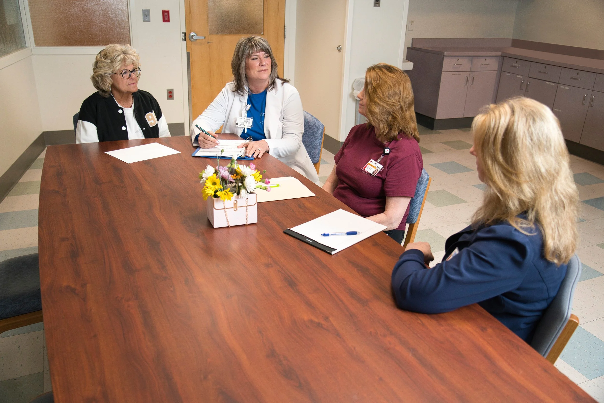 Portrait style photo of four older-aged medical staff women sitting at a wooden table discussing business