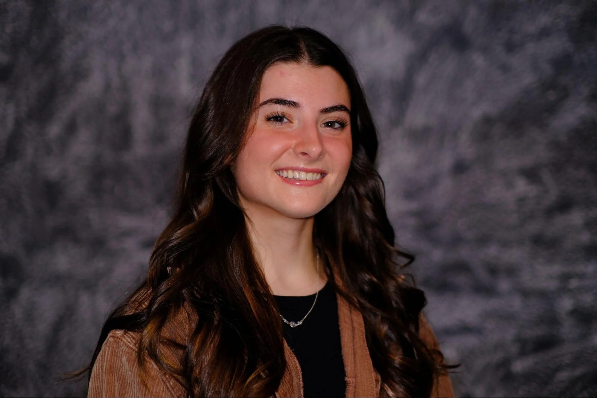 Headshot of a young woman with long dark brown hair wearing a black shirt with a brown overcoat and smiling.