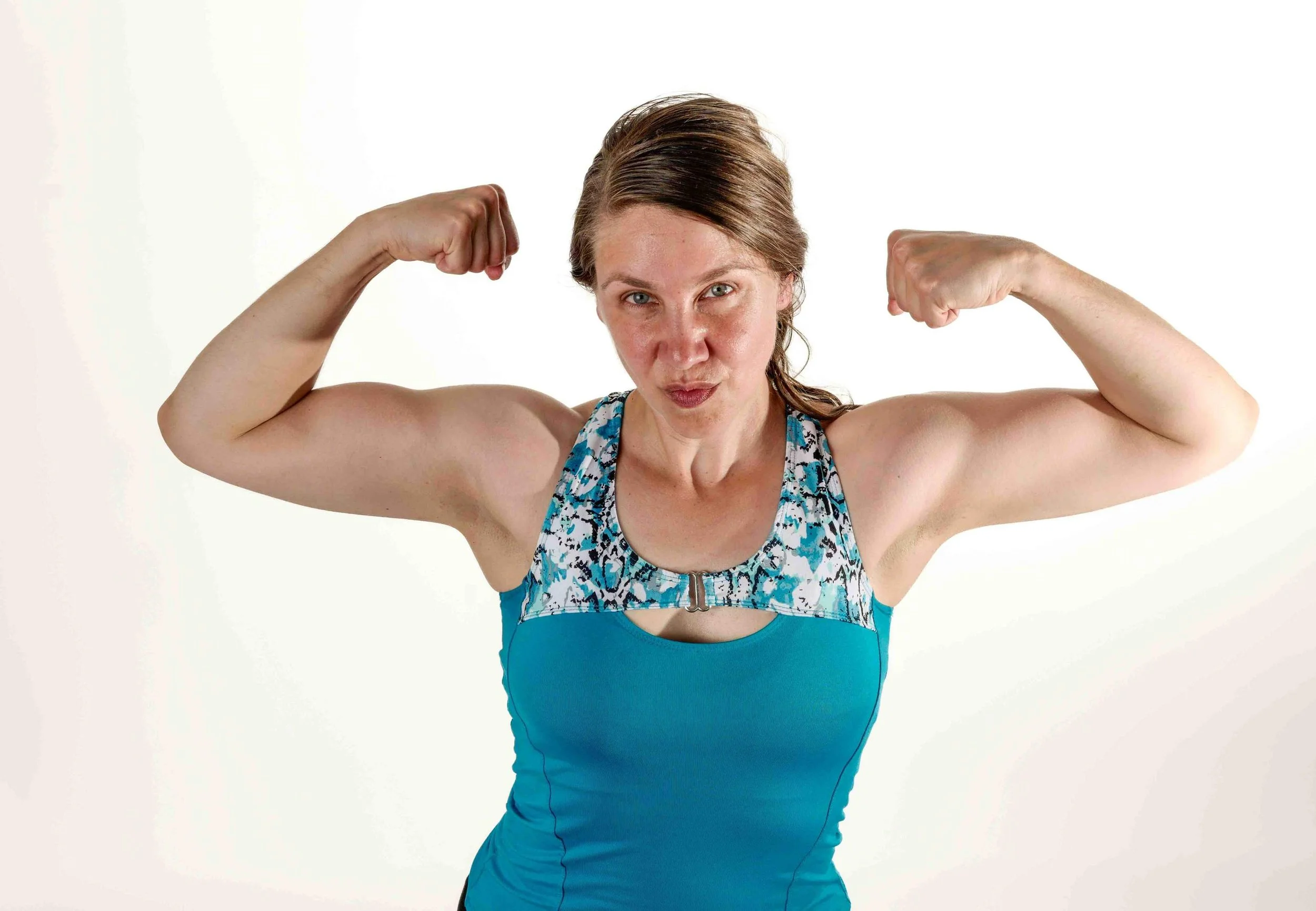 Headshot of a white woman in a blue leotard style top flexing her arm muscles and looking tough
