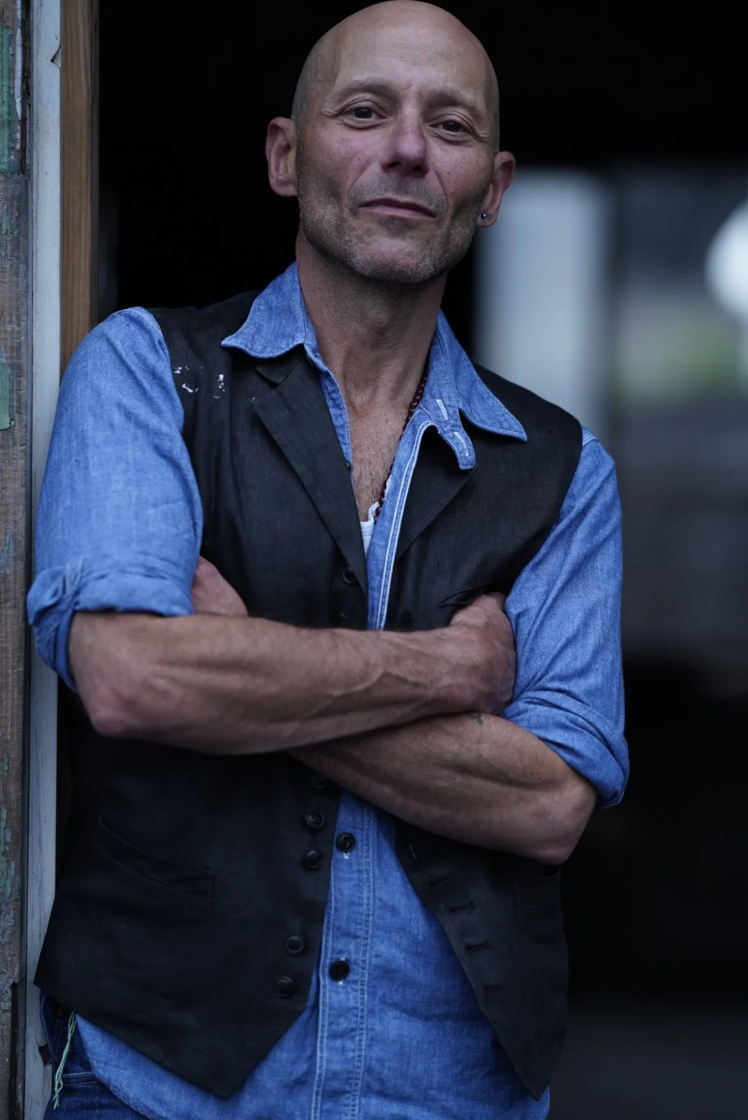 Headshot of a bald man wearing a blue button down with a black vest and arms crossed